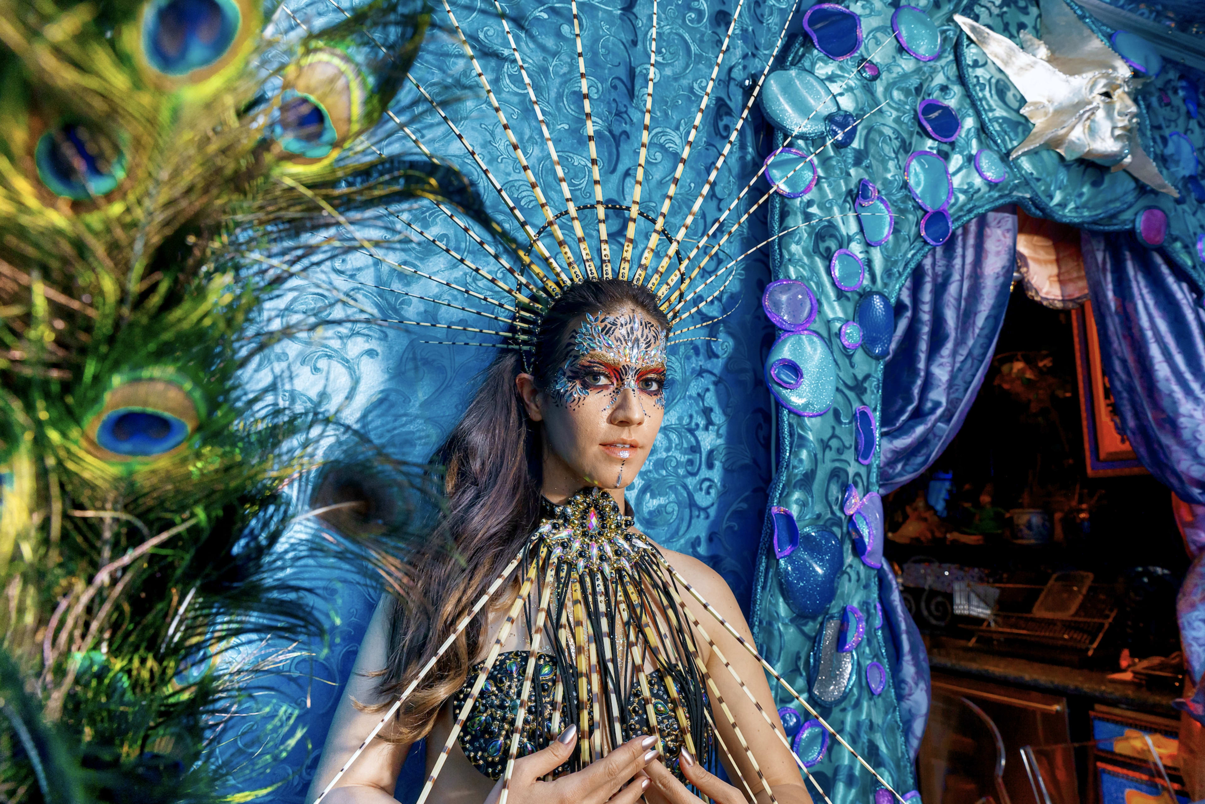 A woman with intricate body art and a jeweled costume stands against a backdrop of textured blue fabrics.