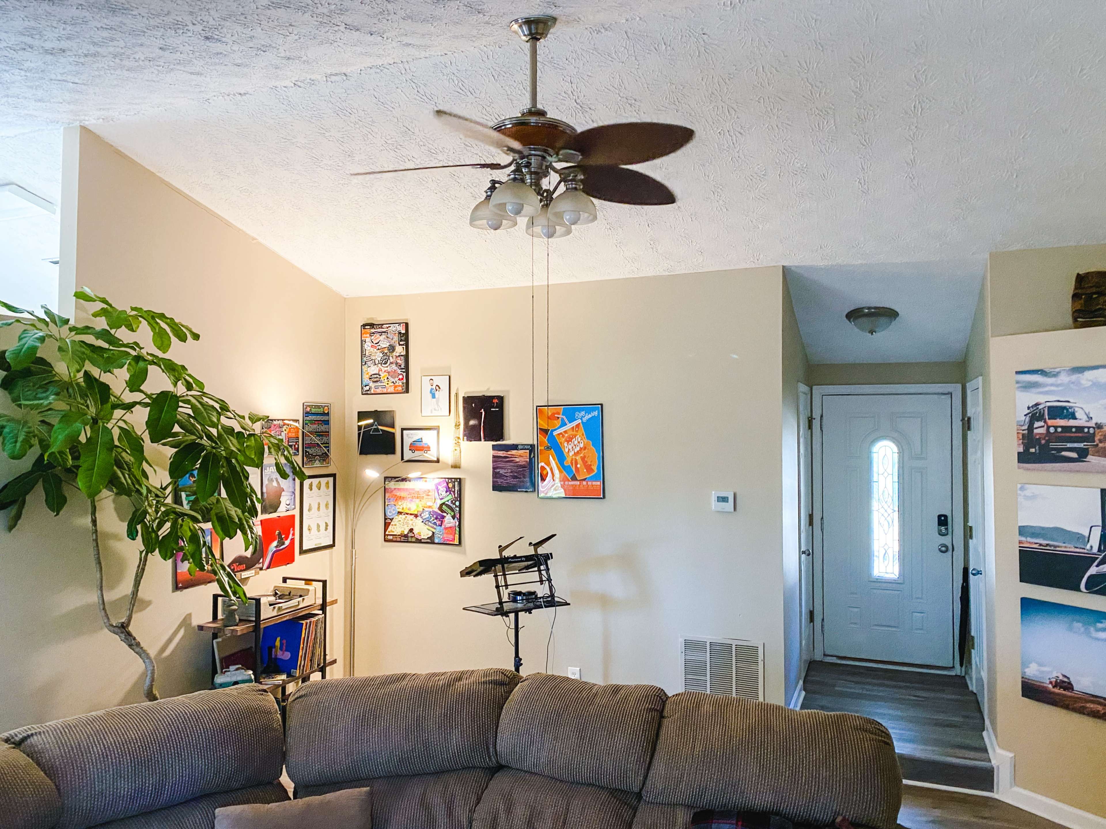 The image shows a living room with a ceiling fan, a large couch, framed artwork on the walls, a potted plant, and a door leading outside.