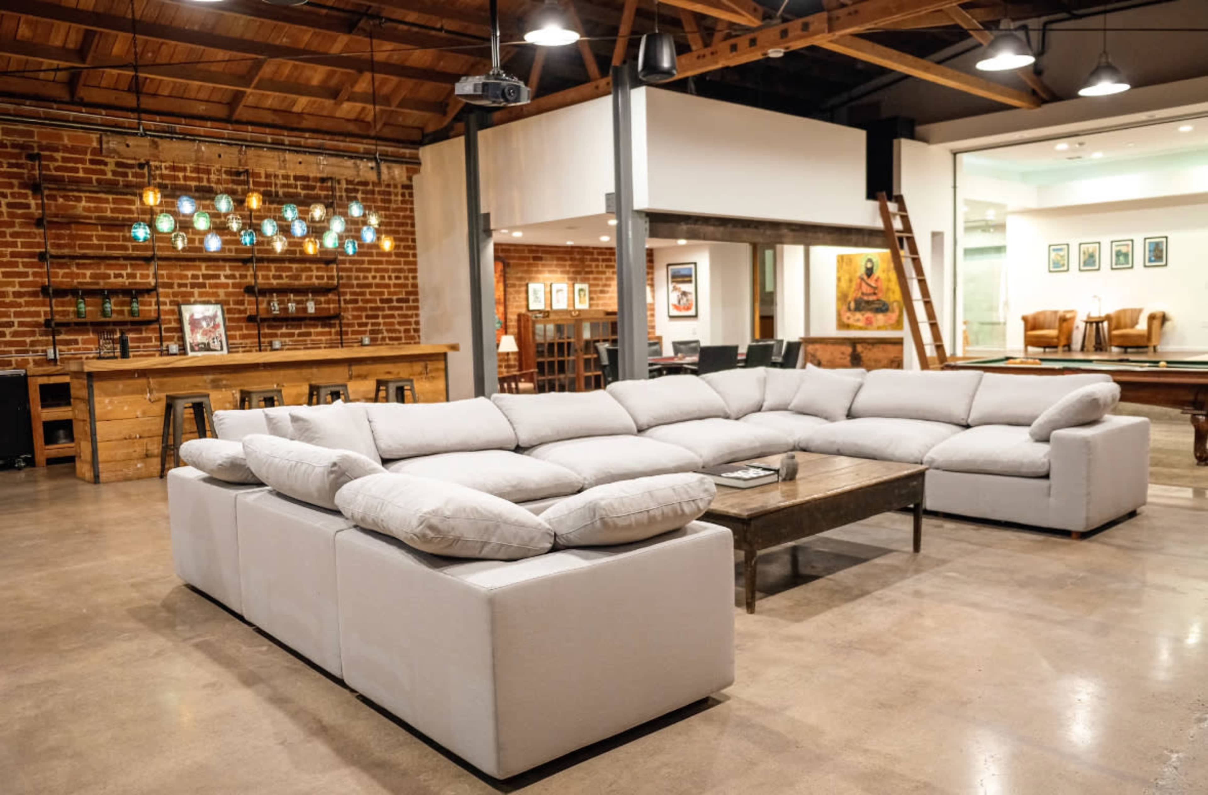 The image shows a spacious, modern lounge area featuring a large sectional sofa, wooden accents, and hanging light fixtures, with a brick wall in the background.