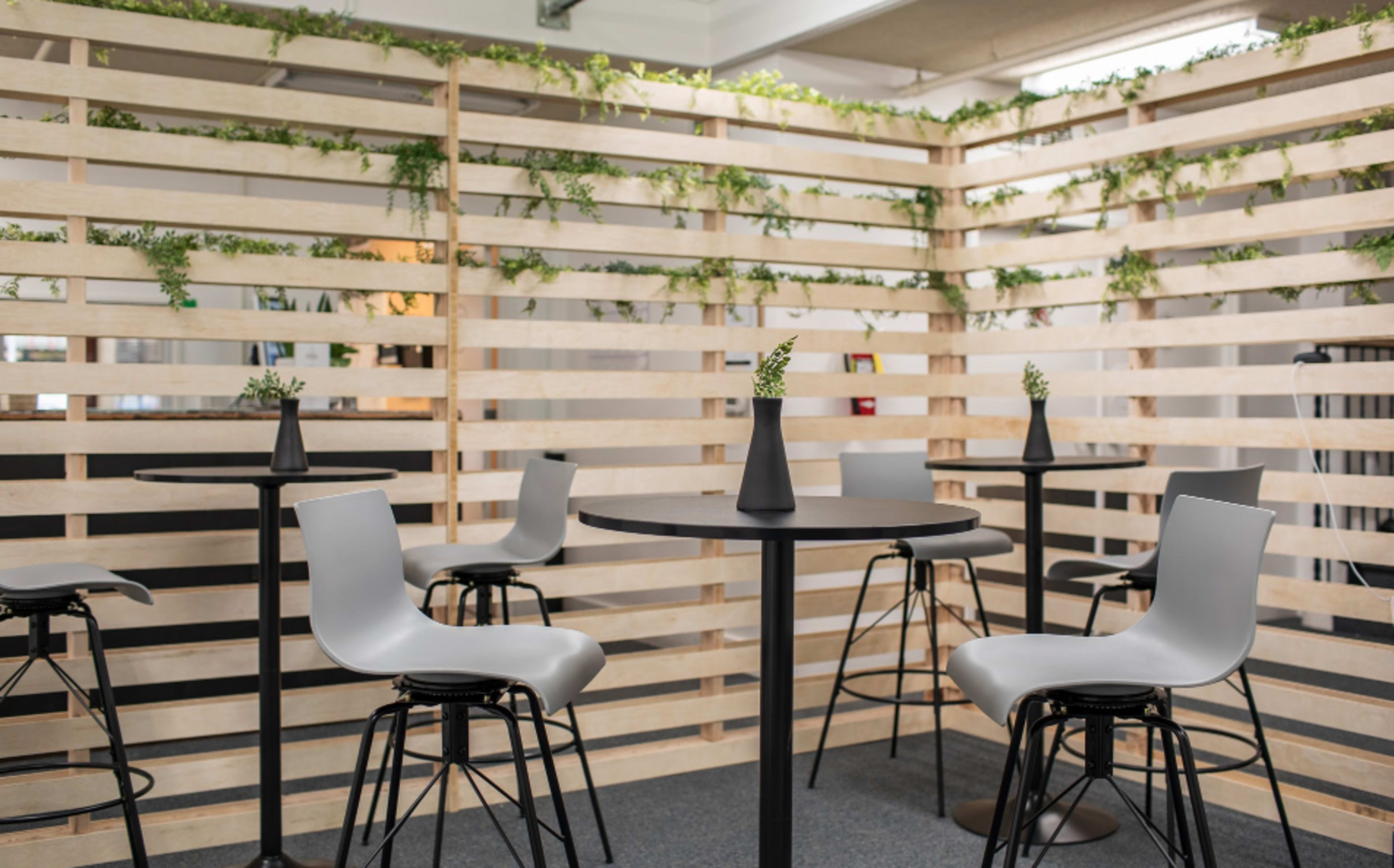 A minimalist seating area with a circular black table and grey chairs surrounded by a wooden slatted divider adorned with greenery.