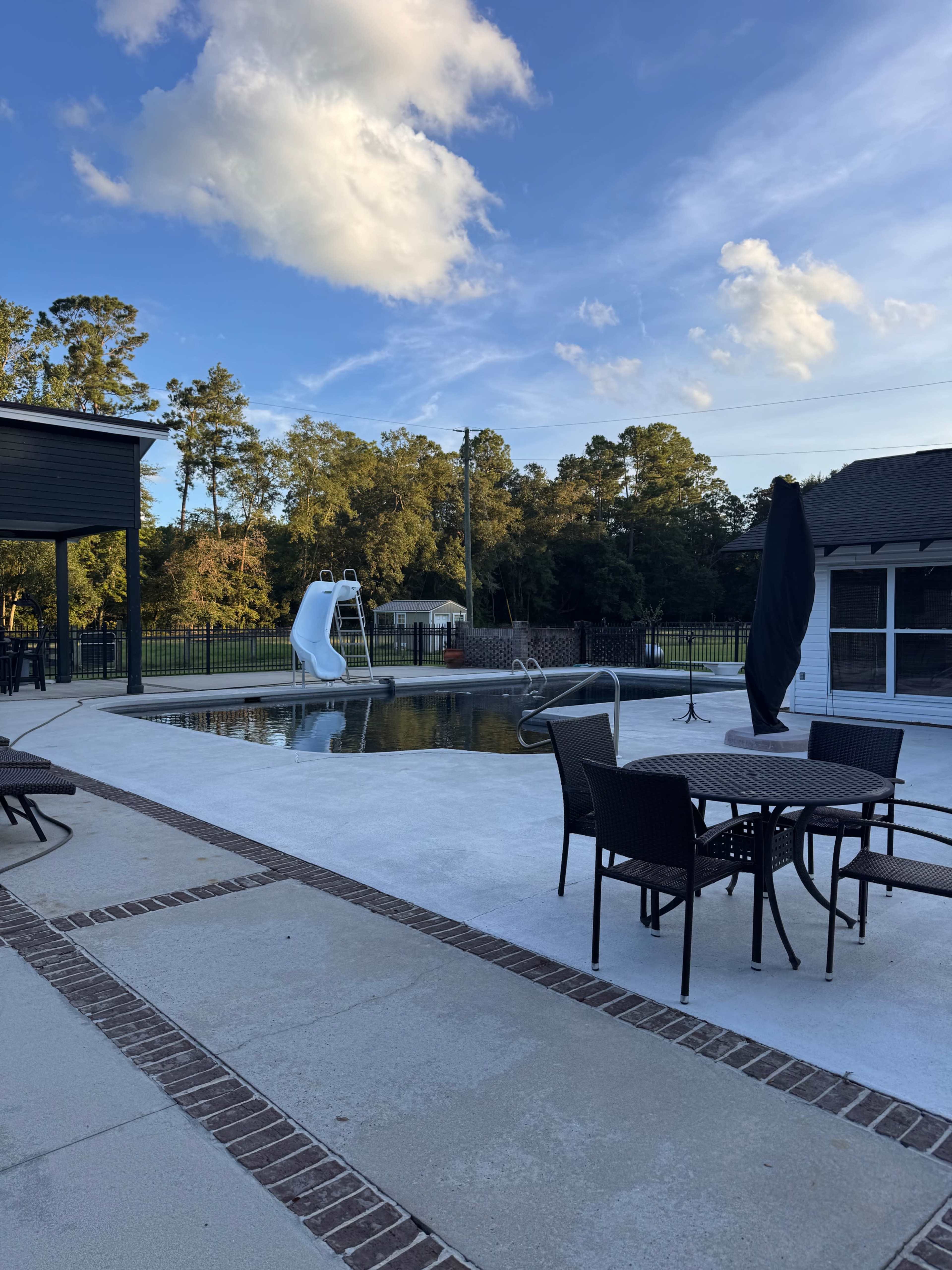 The image shows a swimming pool with a water slide surrounded by patio furniture and a shaded area, set against a backdrop of trees and a clear sky.