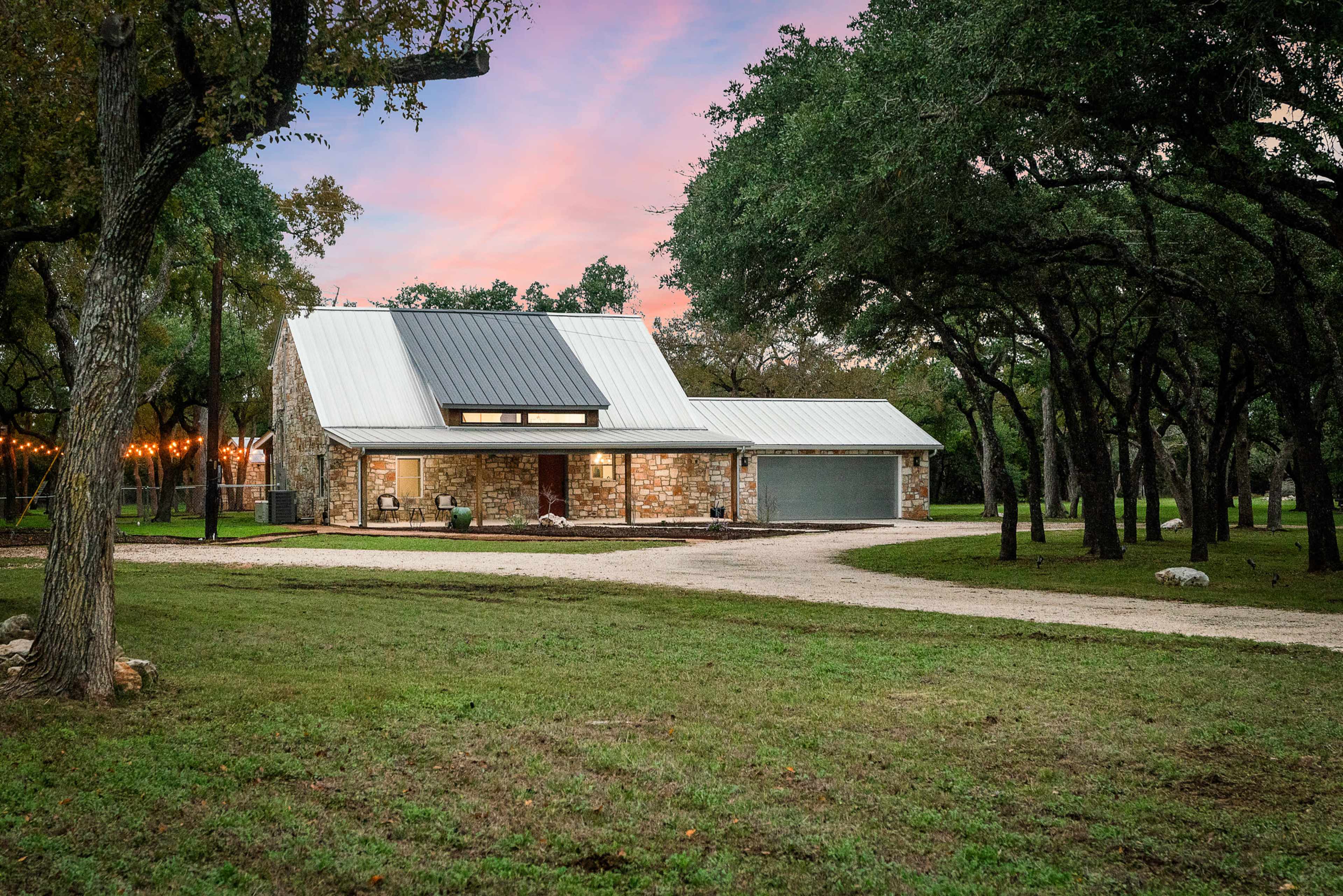 A modern stone house with a metal roof is situated on a gravel driveway surrounded by trees and green grass at dusk.