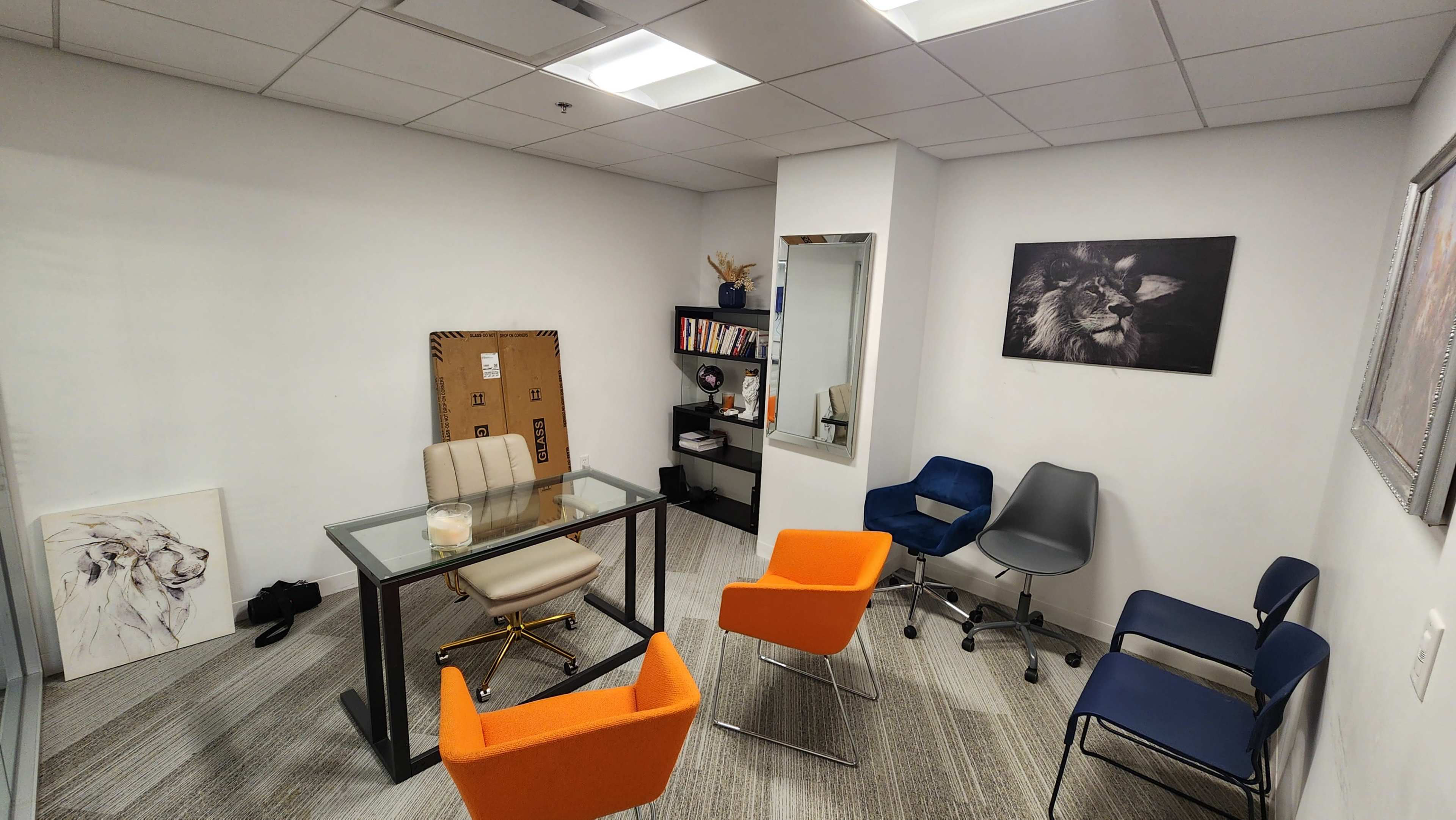 A small office meeting room with a glass table surrounded by orange and blue chairs, a bookcase, and wall art featuring a lion.