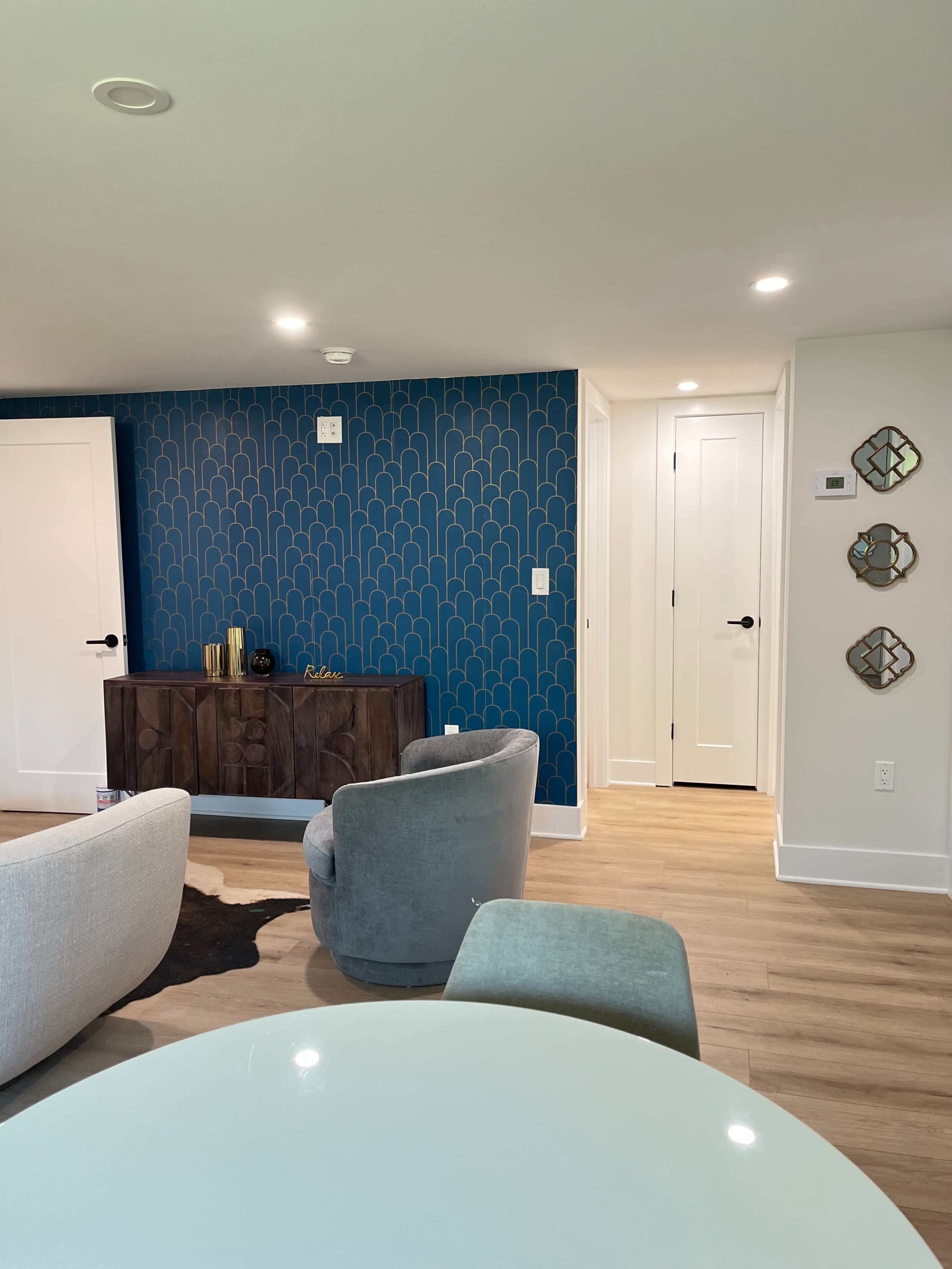 A modern living space featuring a blue scalloped accent wall, a wooden sideboard, and two chairs, with a hallway and doors visible in the background.