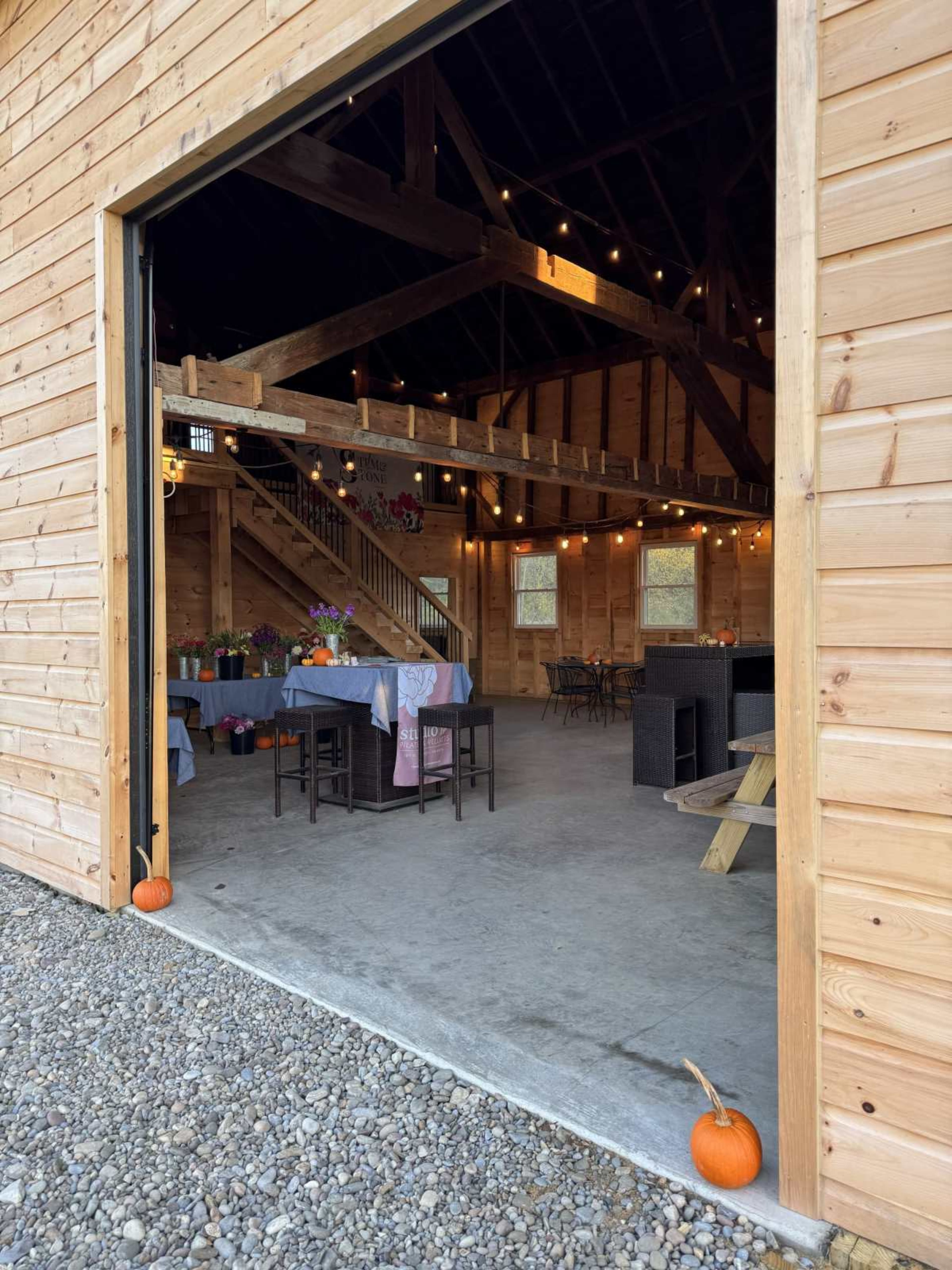 The image shows the interior of a rustic barn with wooden beams, tables set for a gathering, and pumpkins placed at the entrance.