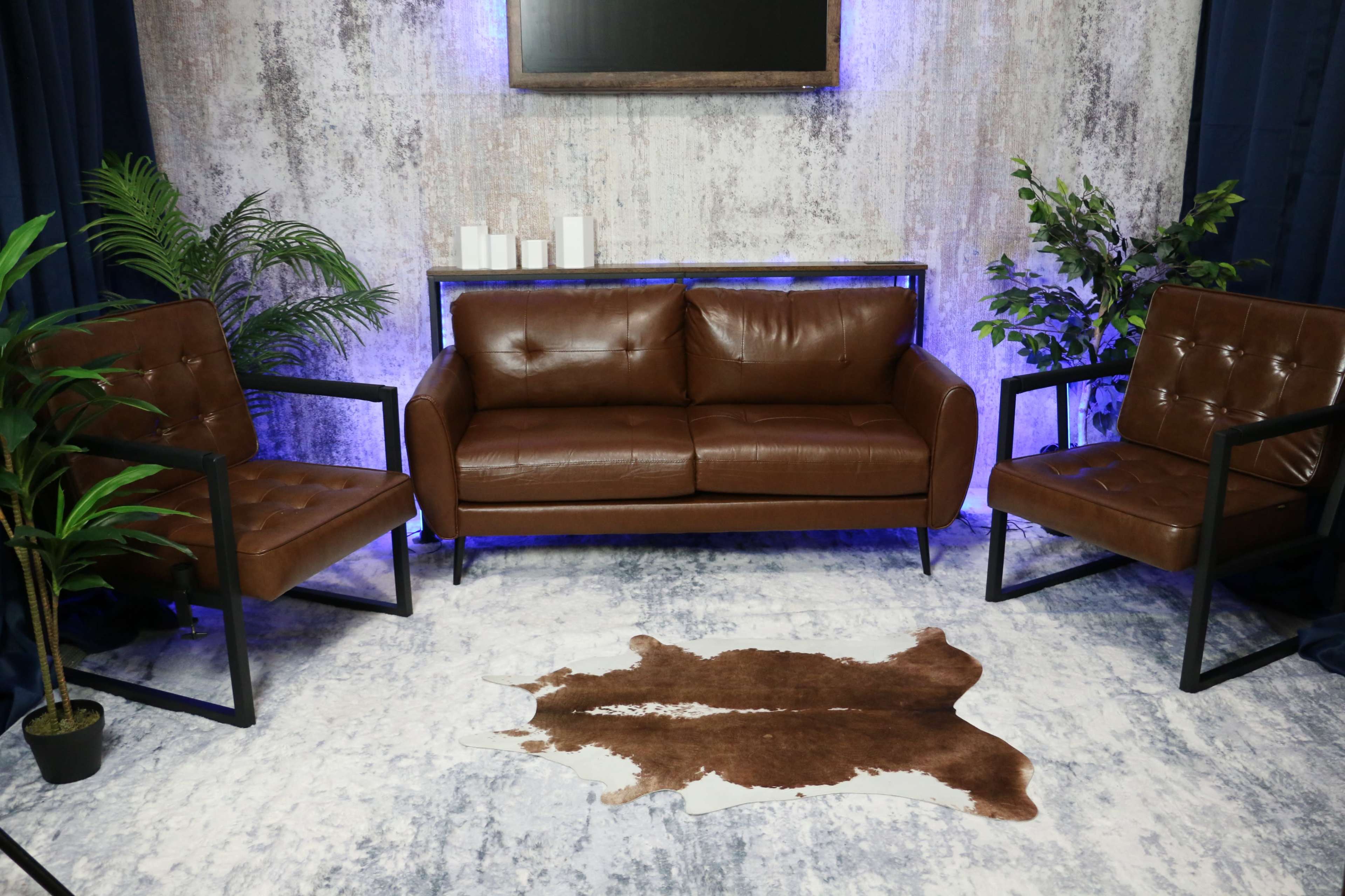 A modern seating area features a brown leather sofa and two matching chairs, set on a patterned rug with a decorative plant and candles nearby.