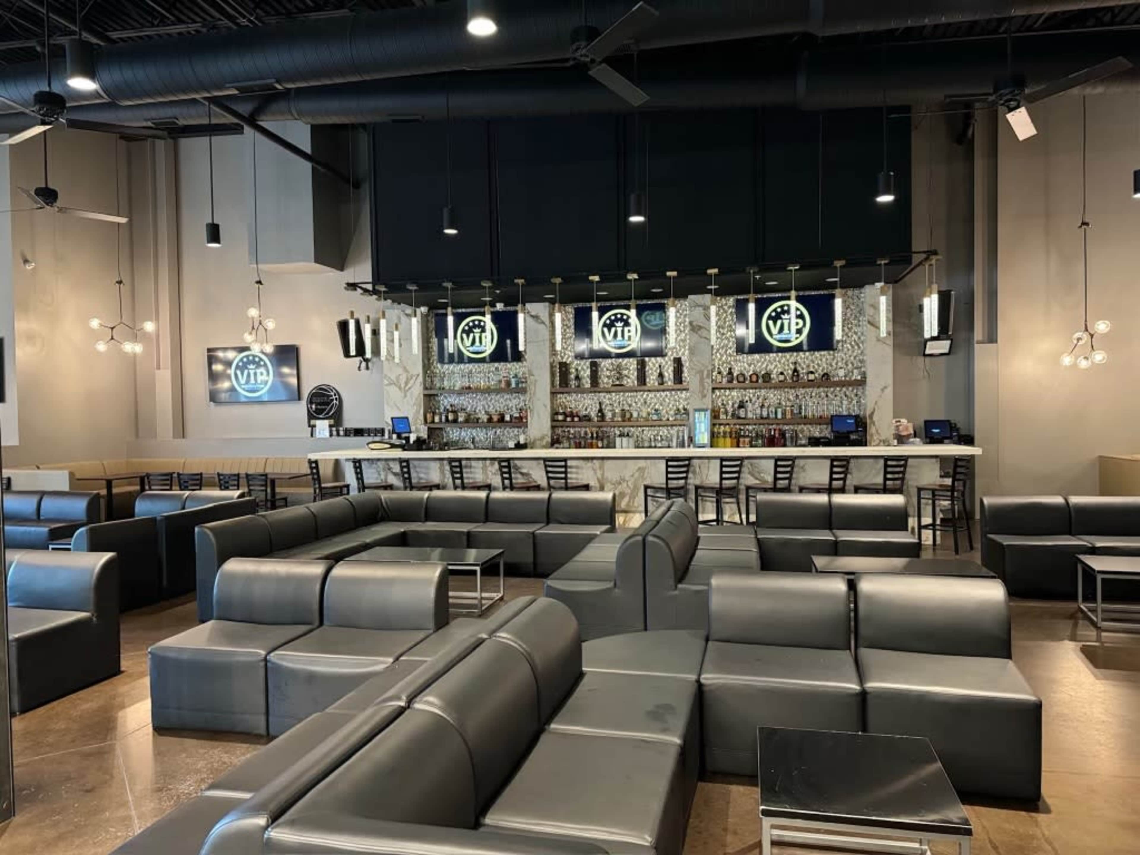A spacious lounge area features black leather seating and a bar with illuminated shelves displaying various bottles.