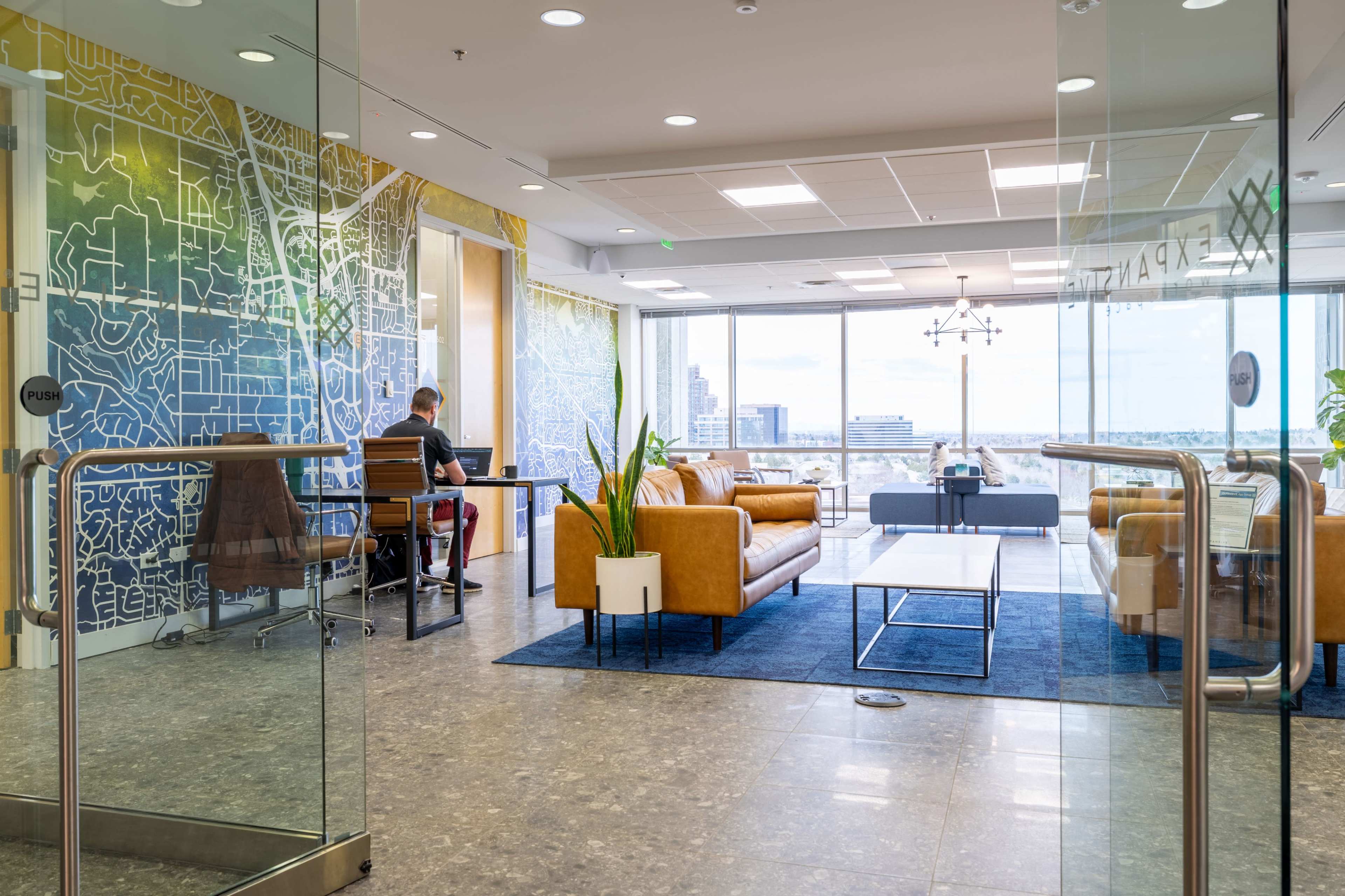 The image shows a modern office space with a person sitting at a table, surrounded by a stylish seating area, large windows, and a colorful map mural on the wall.