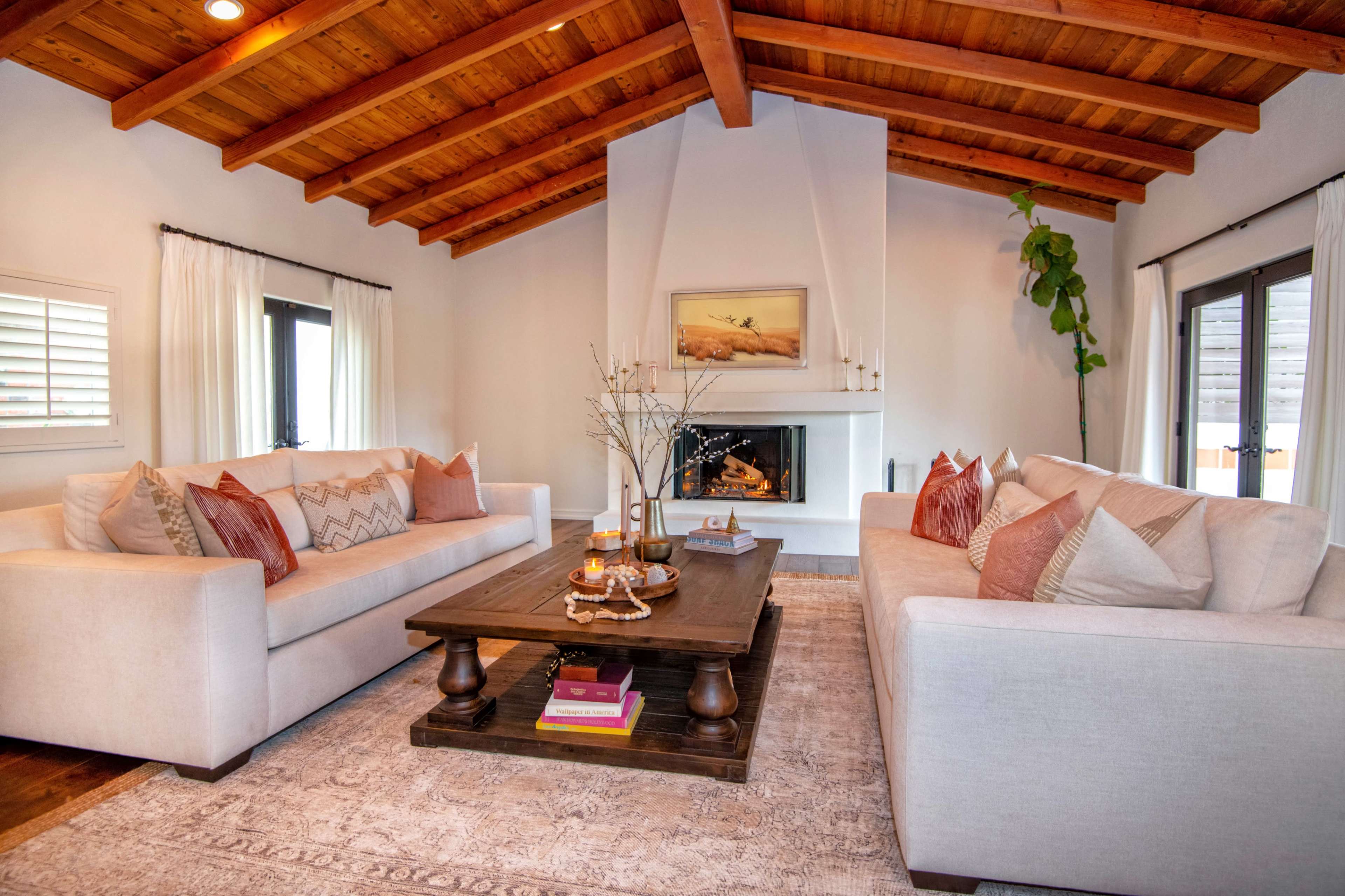 A cozy living room features two sofas facing each other across a wooden coffee table, with a fireplace and a wooden-beamed ceiling in the background.
