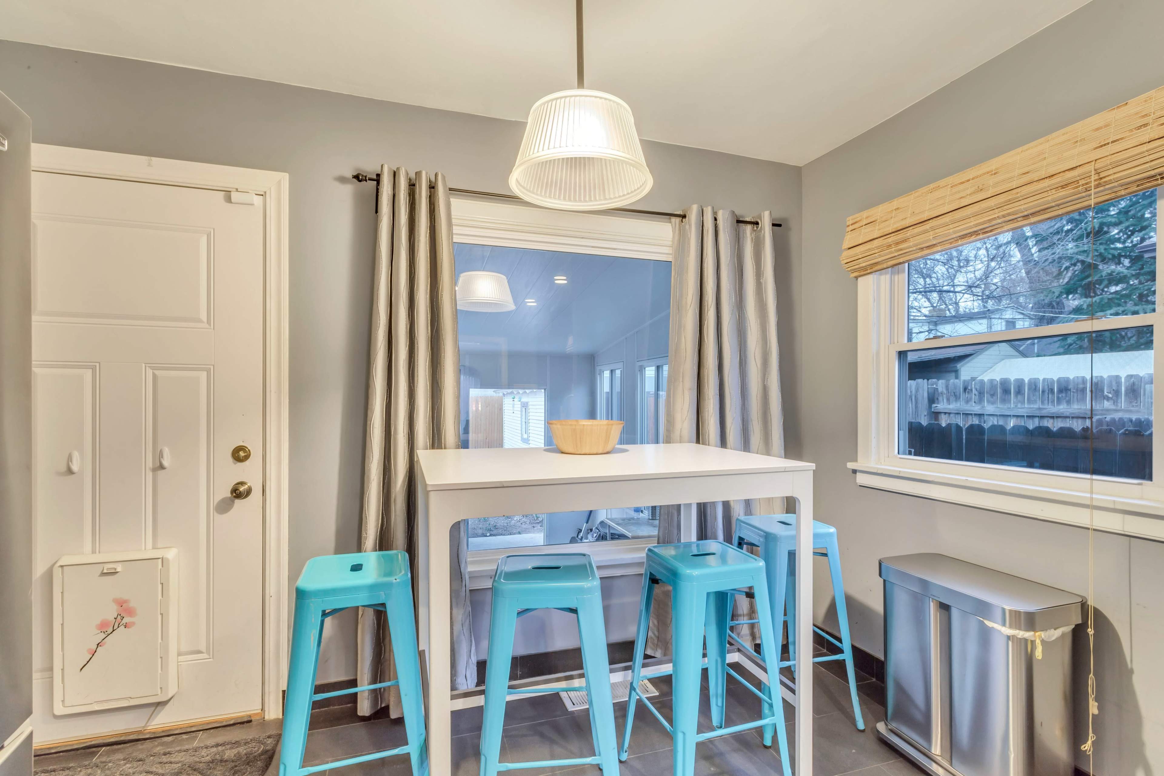 A modern kitchen features a white table surrounded by turquoise stools, with a window dressed in neutral curtains.