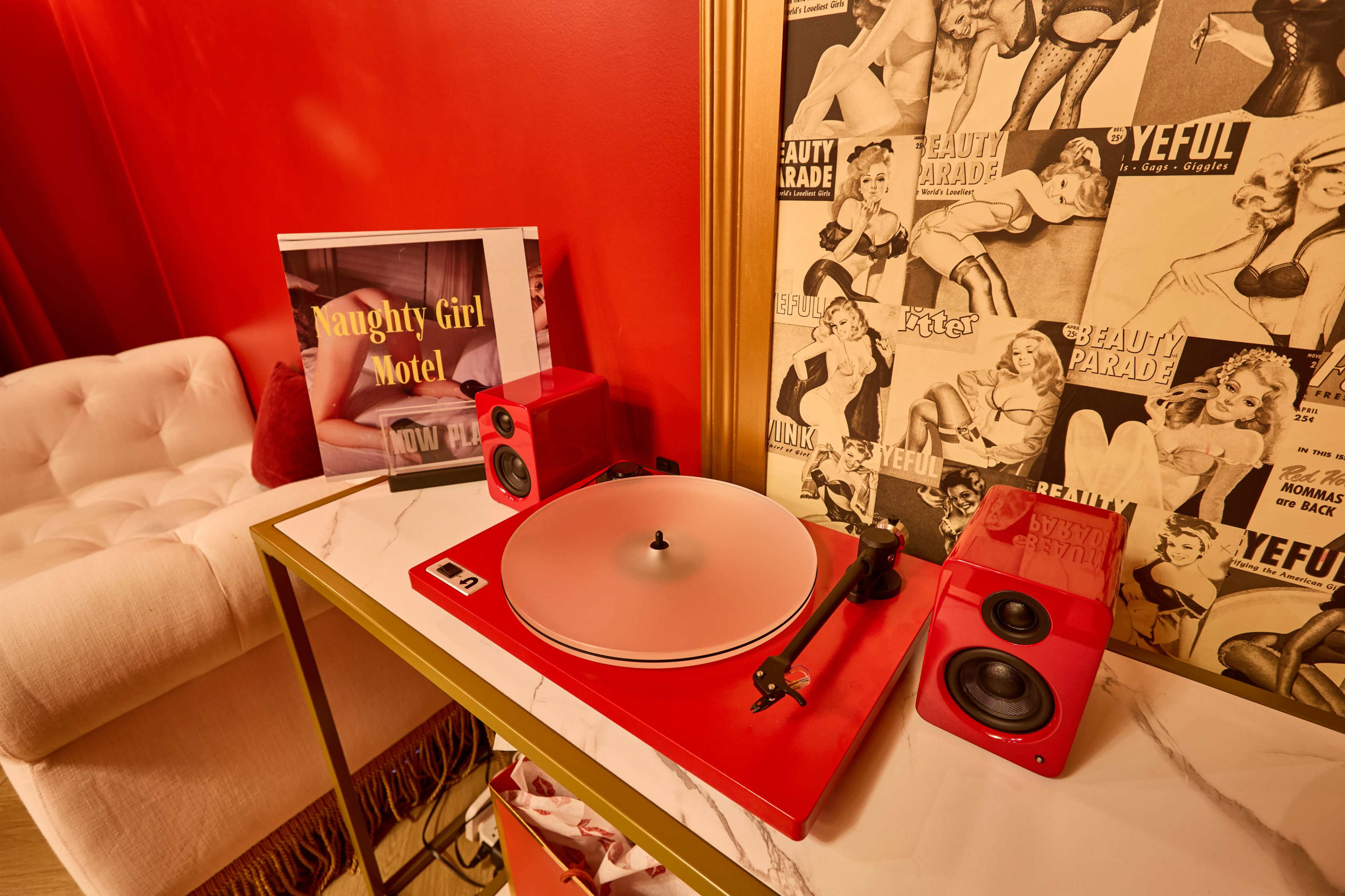 A red record player with matching speakers sits on a marble-topped table beside a framed picture and a wall adorned with vintage pin-up art.