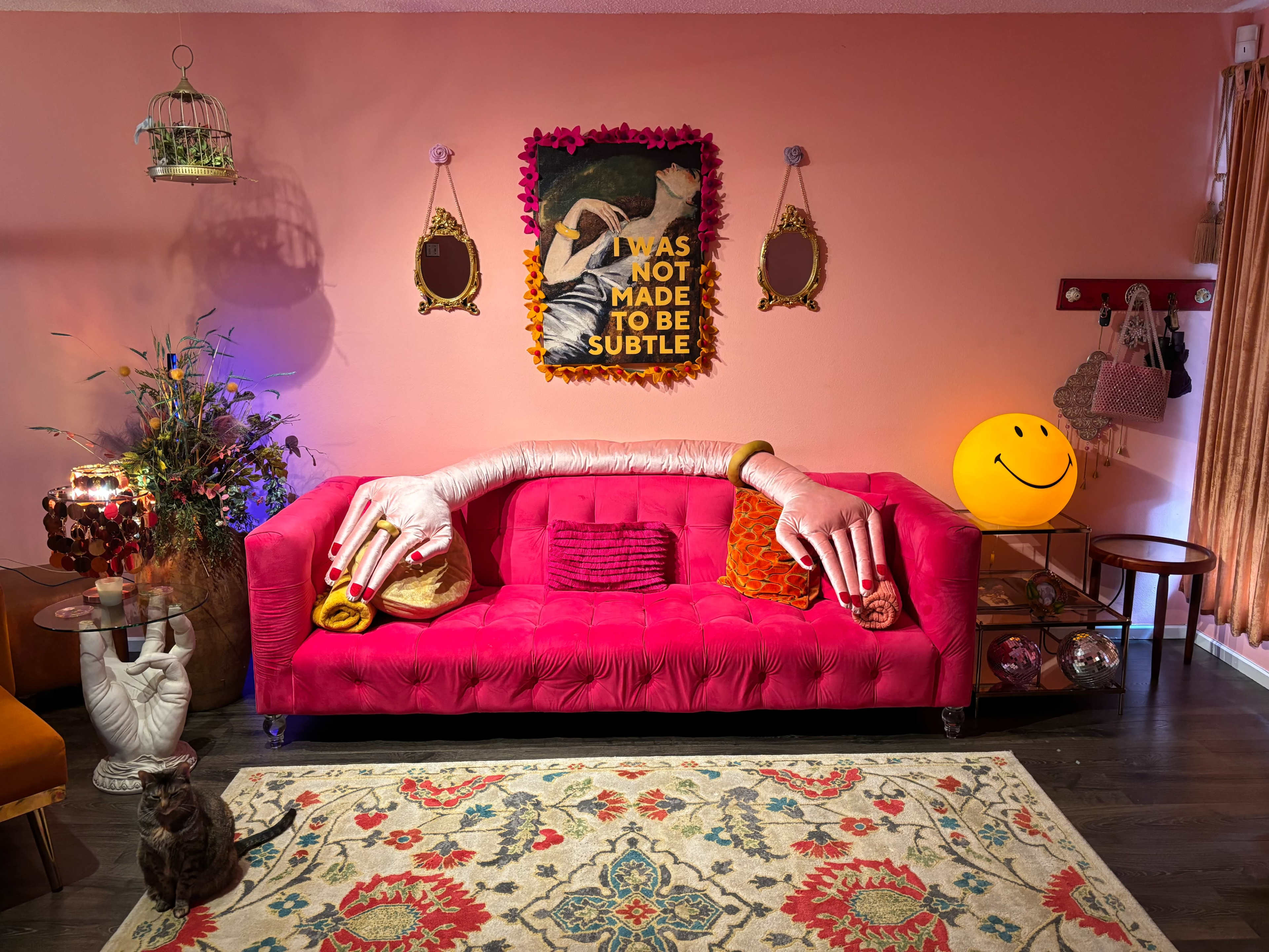 A vibrant pink couch with oversized decorative hands rests against a peach wall, surrounded by eclectic decor, including mirrors, a smiling lamp, and a colorful rug.