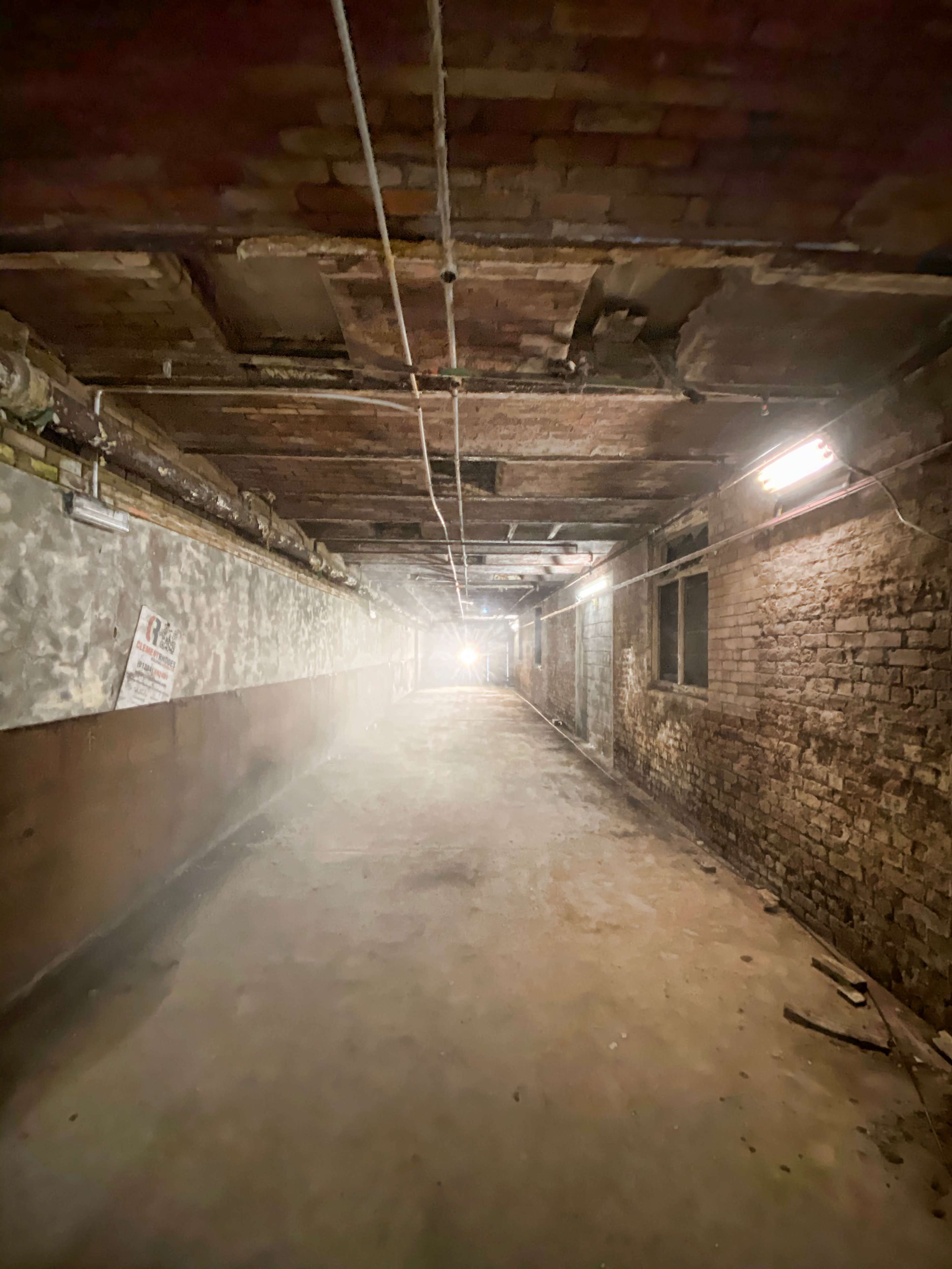 A dimly lit underground corridor with exposed brick walls and a cracked ceiling leads to a bright light at the far end.