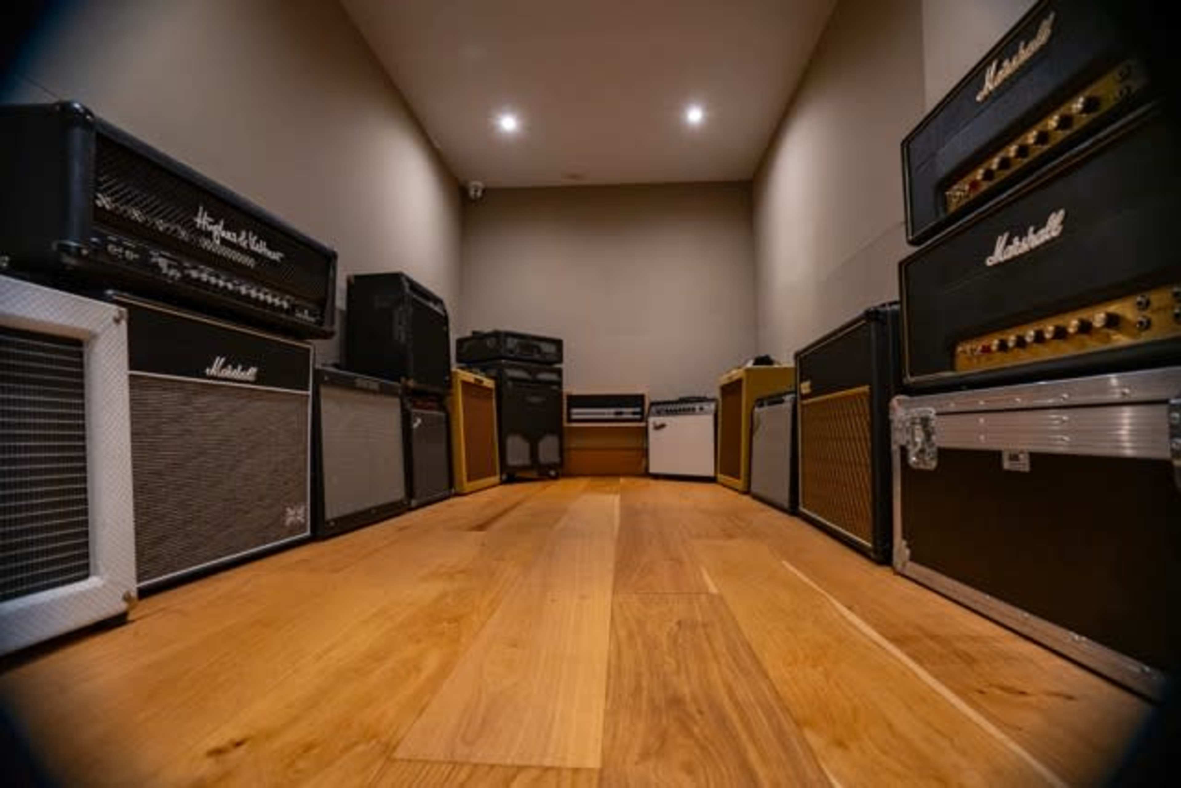 The image features a room filled with various guitar amplifiers arranged along the walls, with wooden flooring and soft lighting.