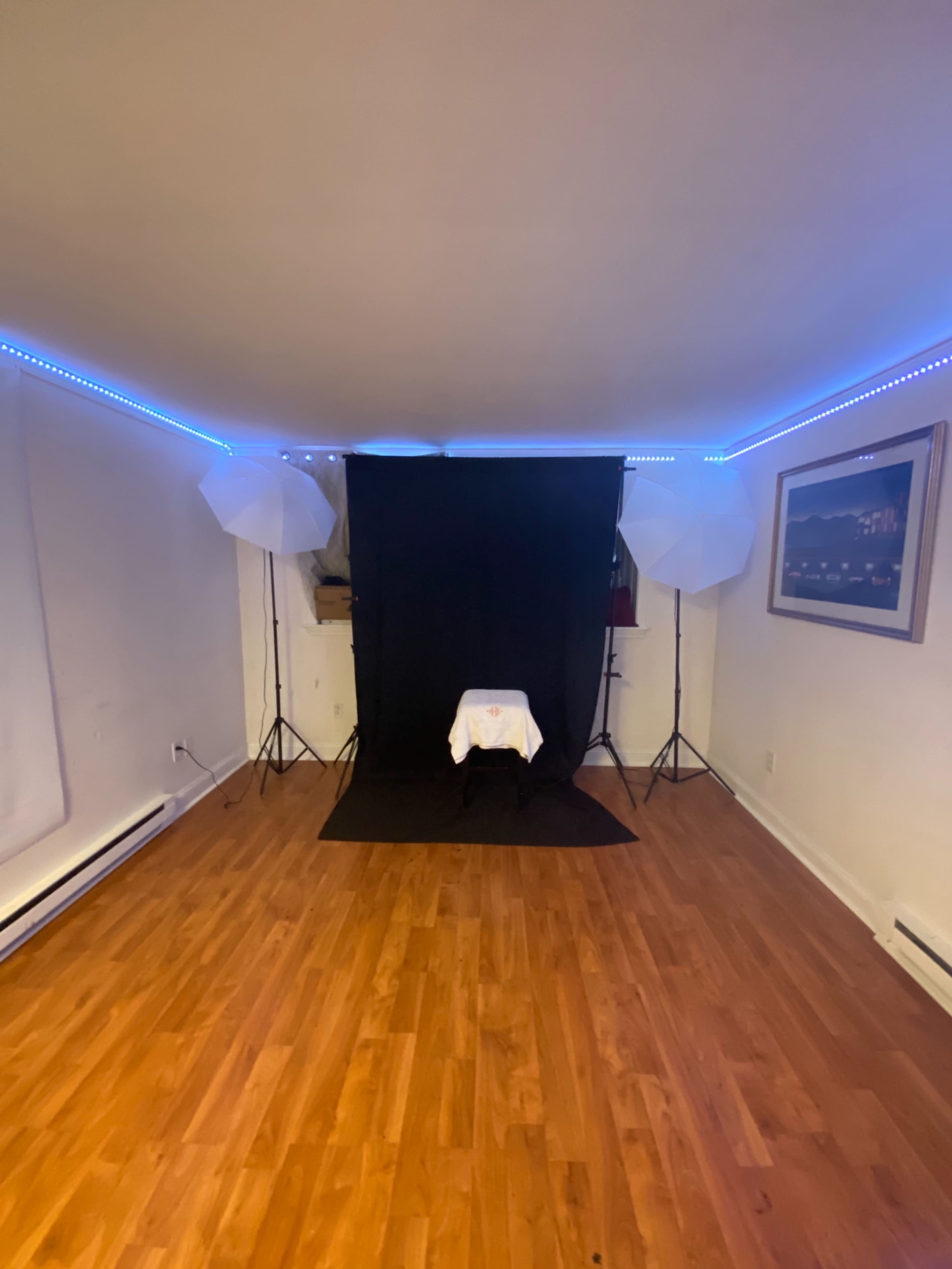 The image shows a photography studio setup with a black backdrop, two light stands with umbrellas, and a table covered with a white cloth in a room with wooden flooring.