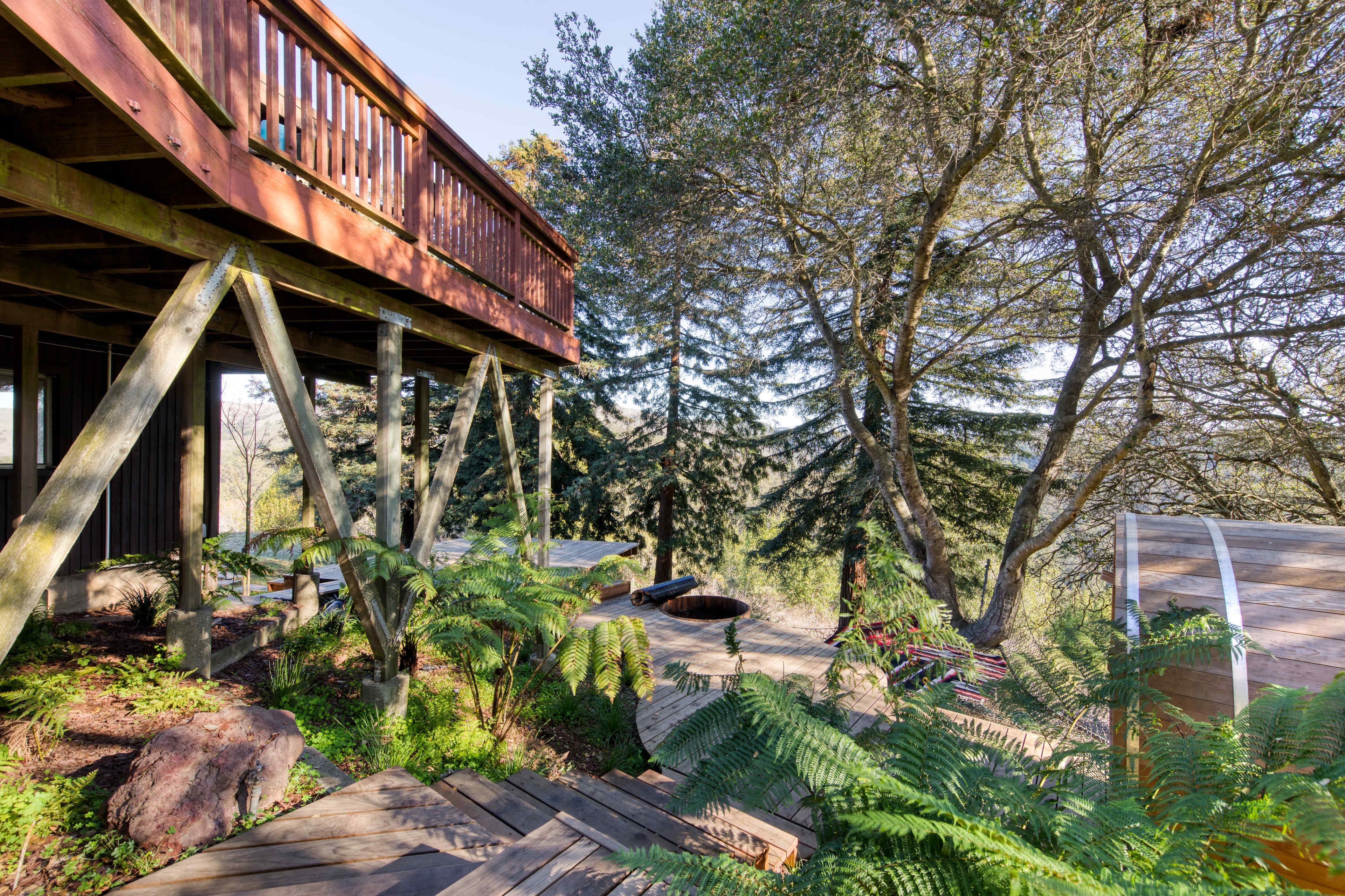 A wooden deck elevated above a garden area with ferns and trees, featuring a circular stone fire pit and a winding path made of wooden planks.