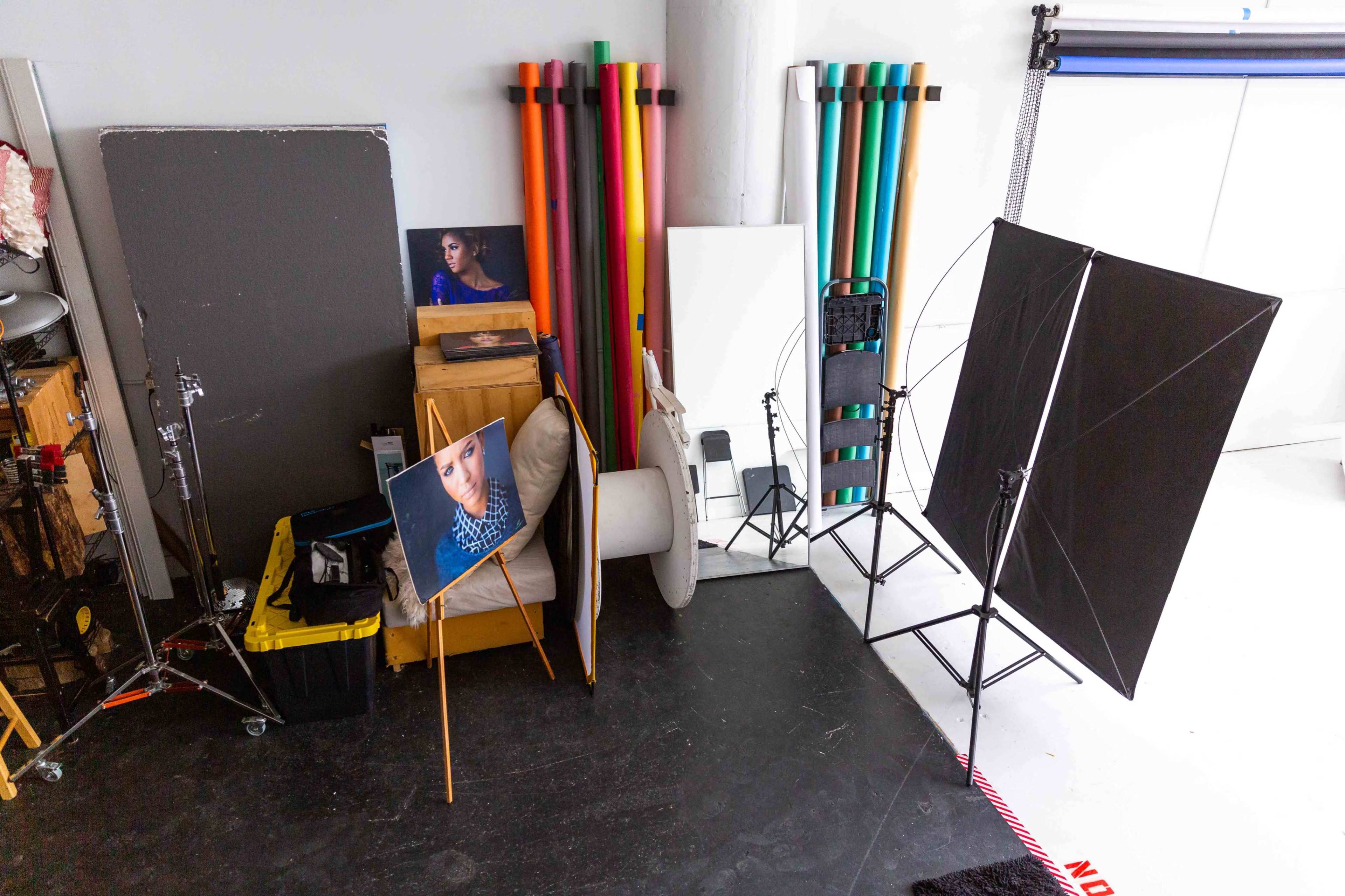 The image shows a photography studio with a corner setup featuring a portrait on an easel, backdrop rolls, lighting equipment, and various studio props.