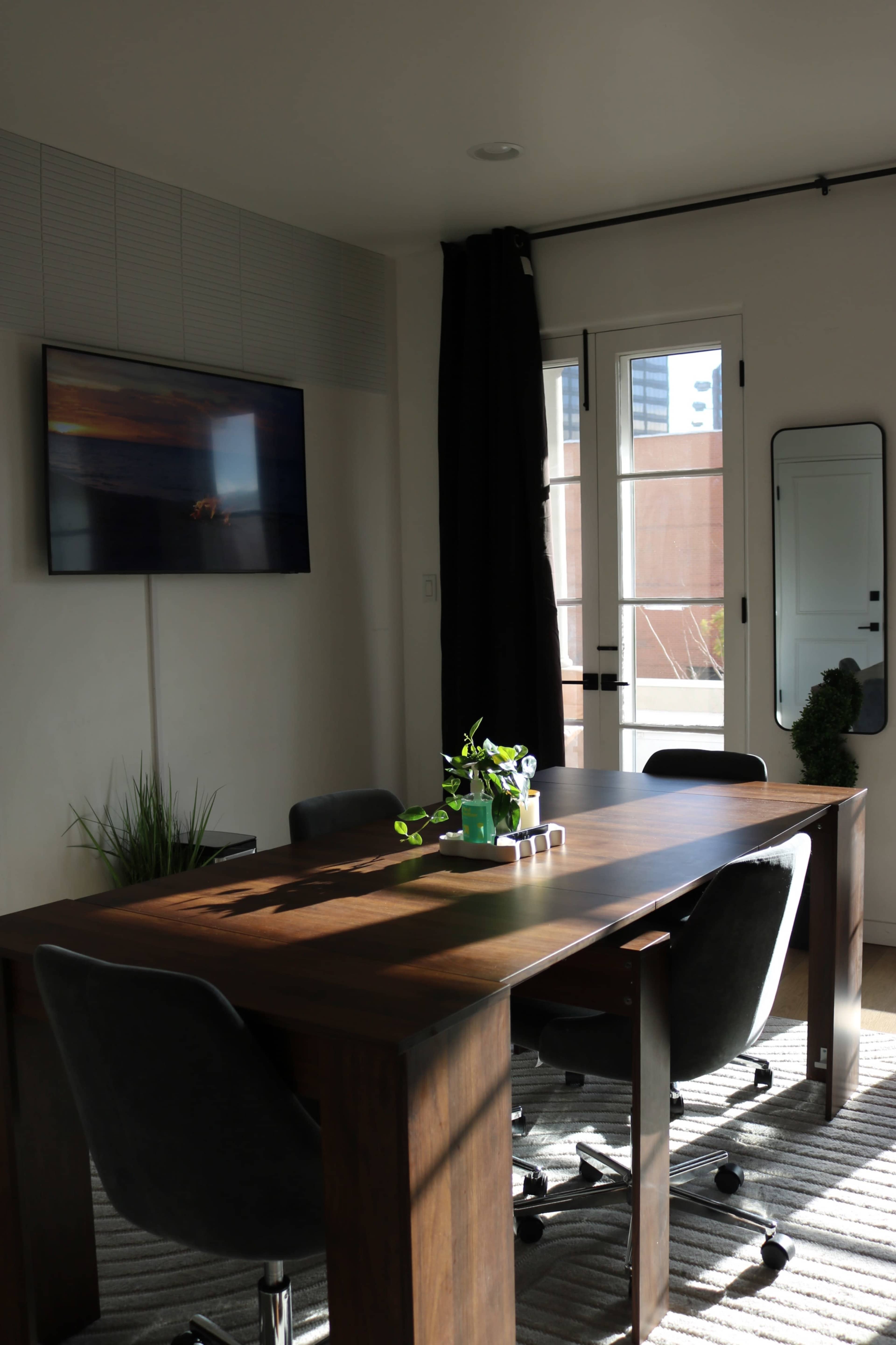 The image shows a modern meeting room with a wooden table, black chairs, a television mounted on the wall, and a large window leading to a balcony.