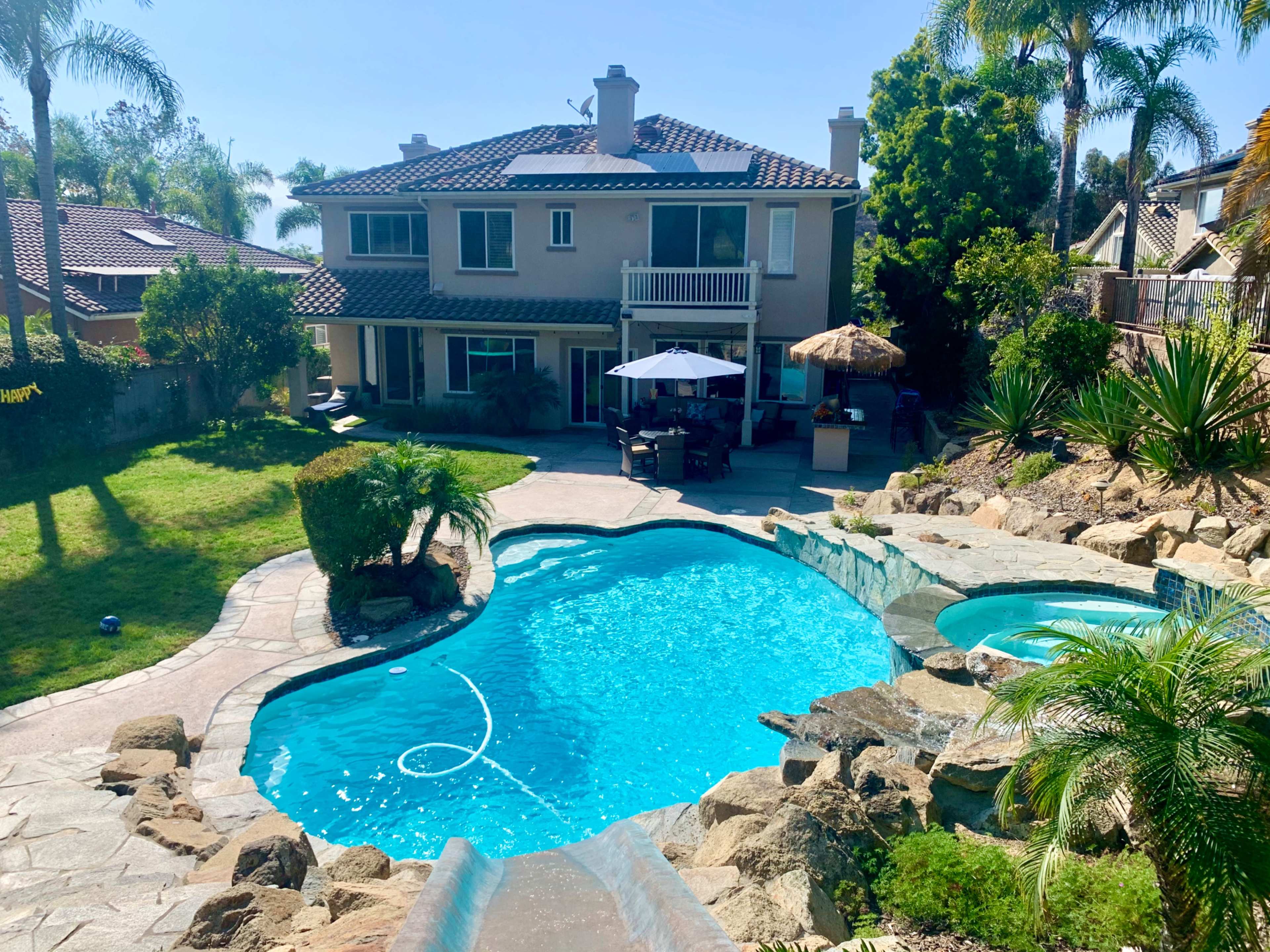 A large backyard featuring a landscaped swimming pool with a rock slide, surrounded by palm trees and a two-story house in the background.