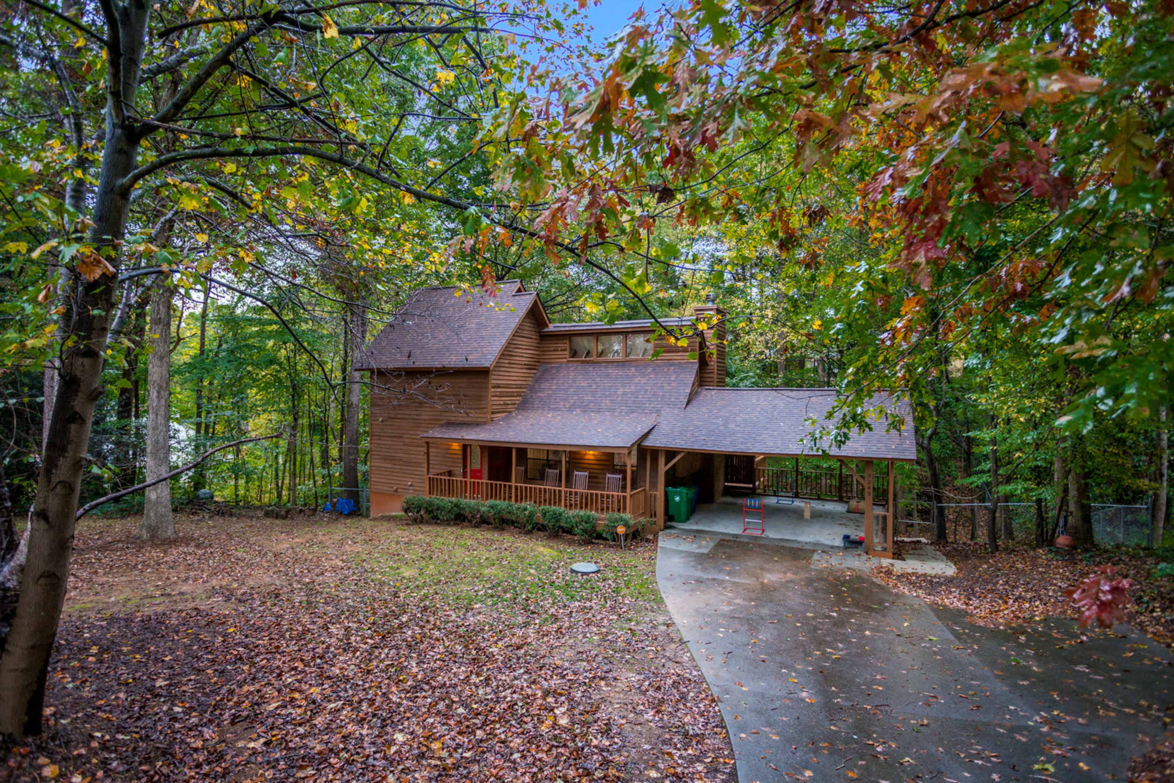 A wooden house surrounded by trees, with a covered porch and a leaf-strewn driveway.