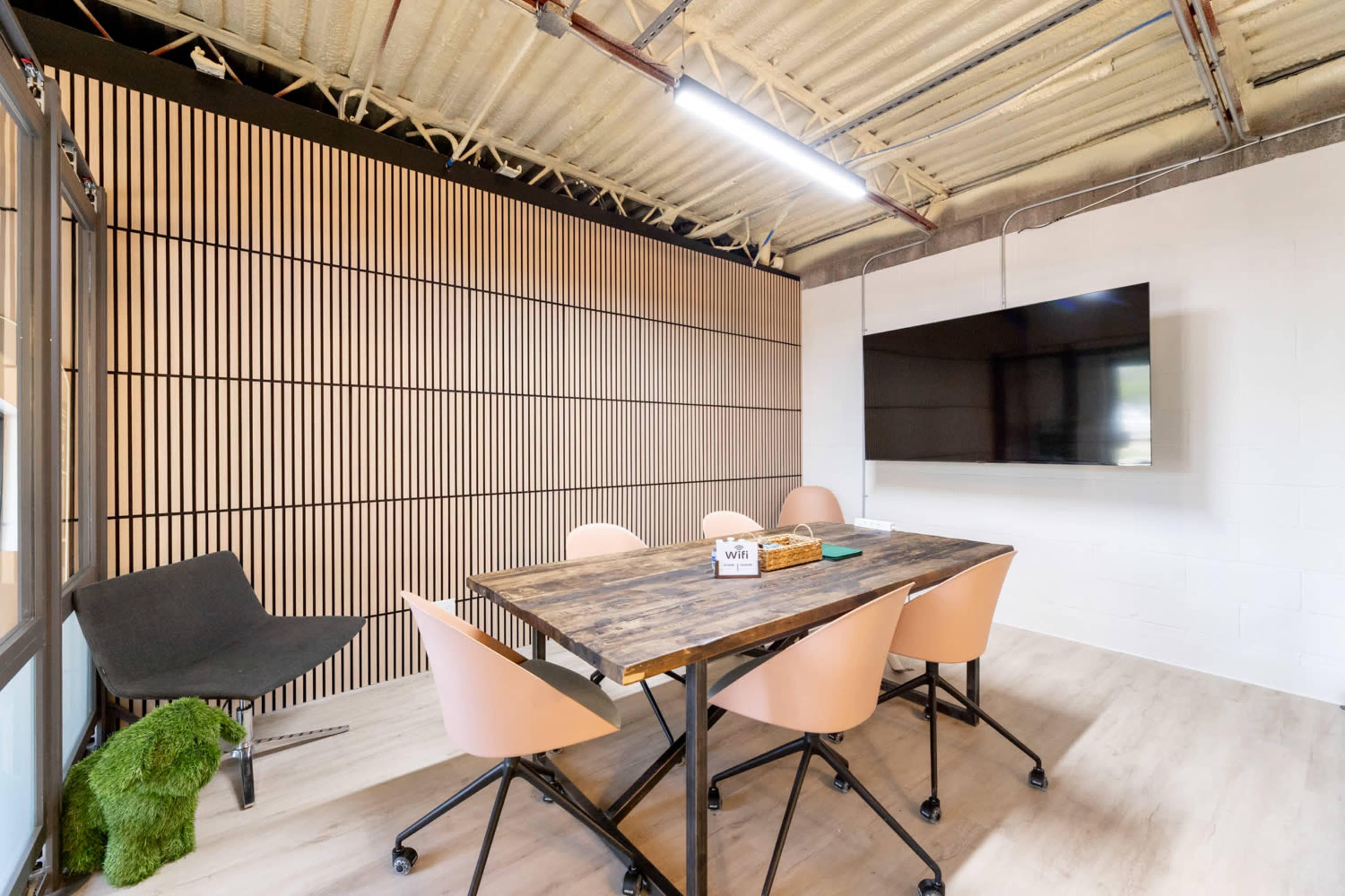 The image shows a modern meeting room with a wooden table surrounded by pink chairs, a television mounted on the wall, and a striped wooden panel backdrop.