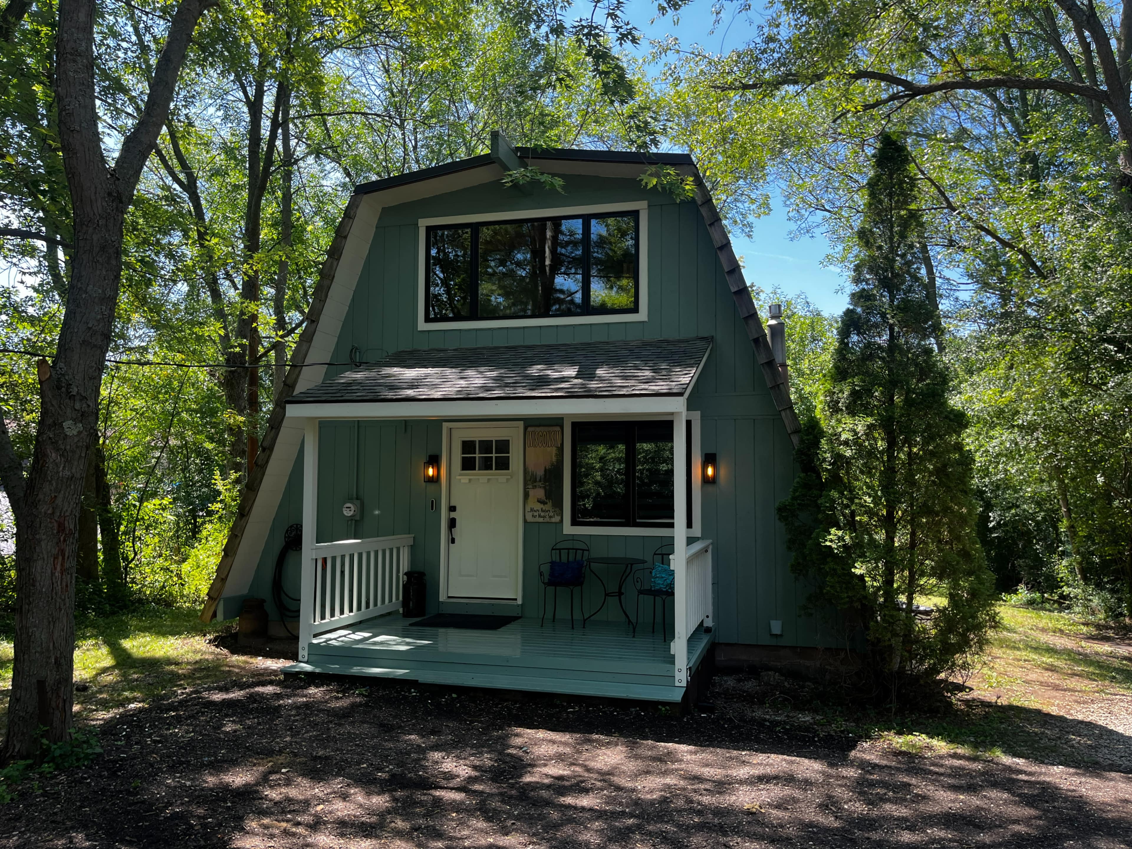 A light blue, A-frame house is surrounded by trees, featuring a front porch with two chairs and a small table.