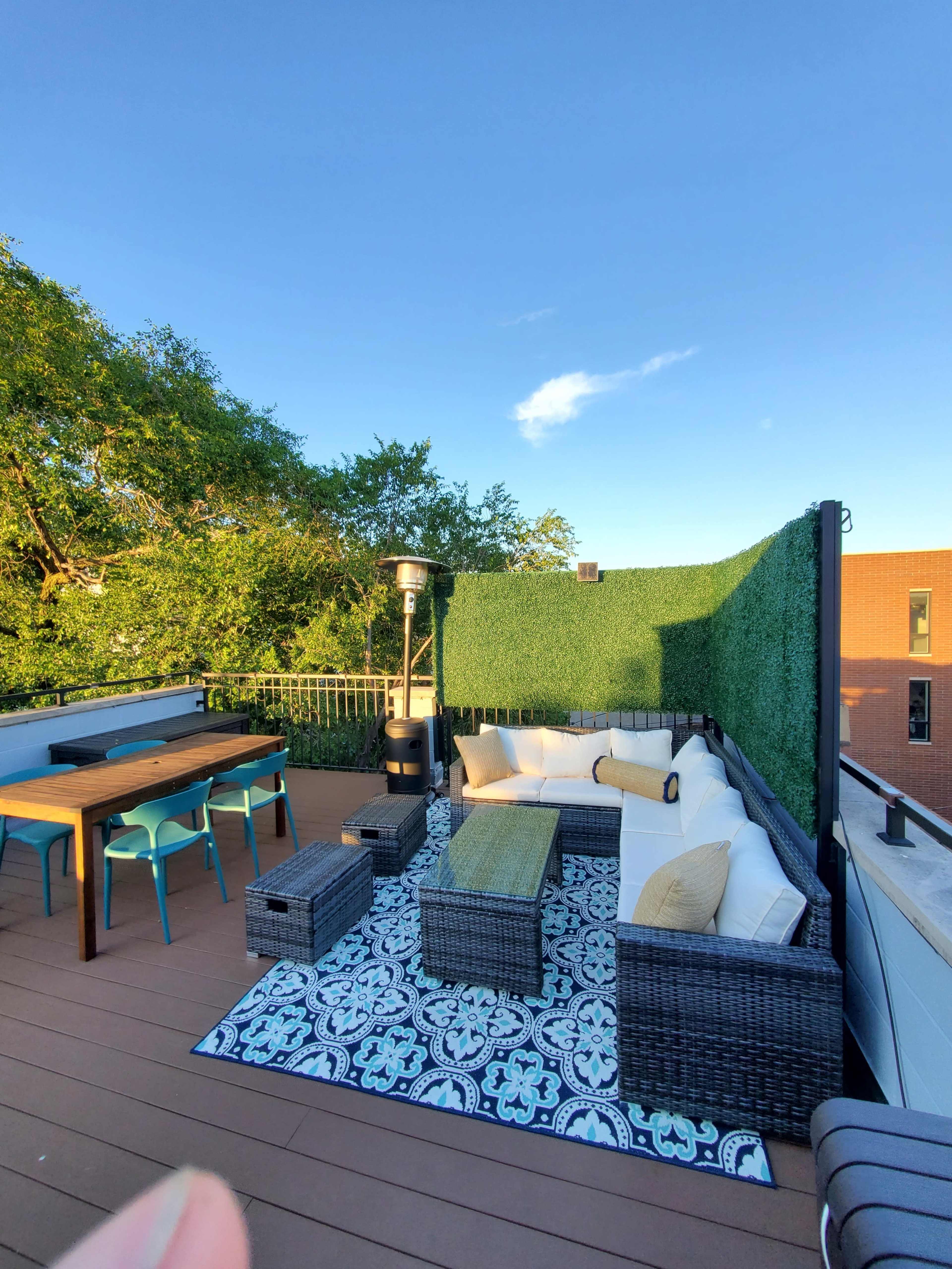 A rooftop terrace features a seating area with wicker furniture, a dining table, and green foliage along the perimeter under a clear blue sky.
