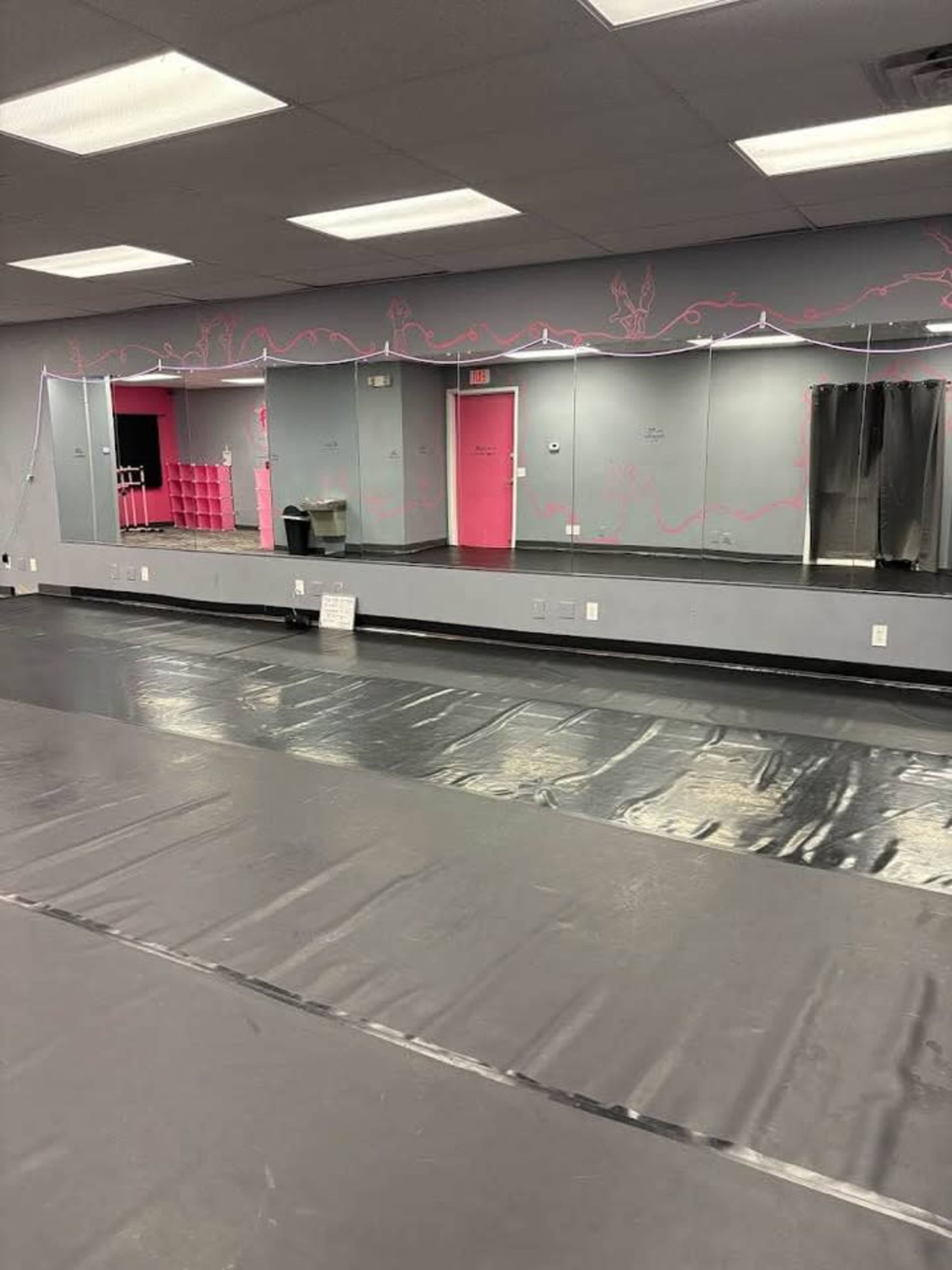 The image shows an empty studio with a black floor, mirrored walls, and pink accent elements, including a storage unit.