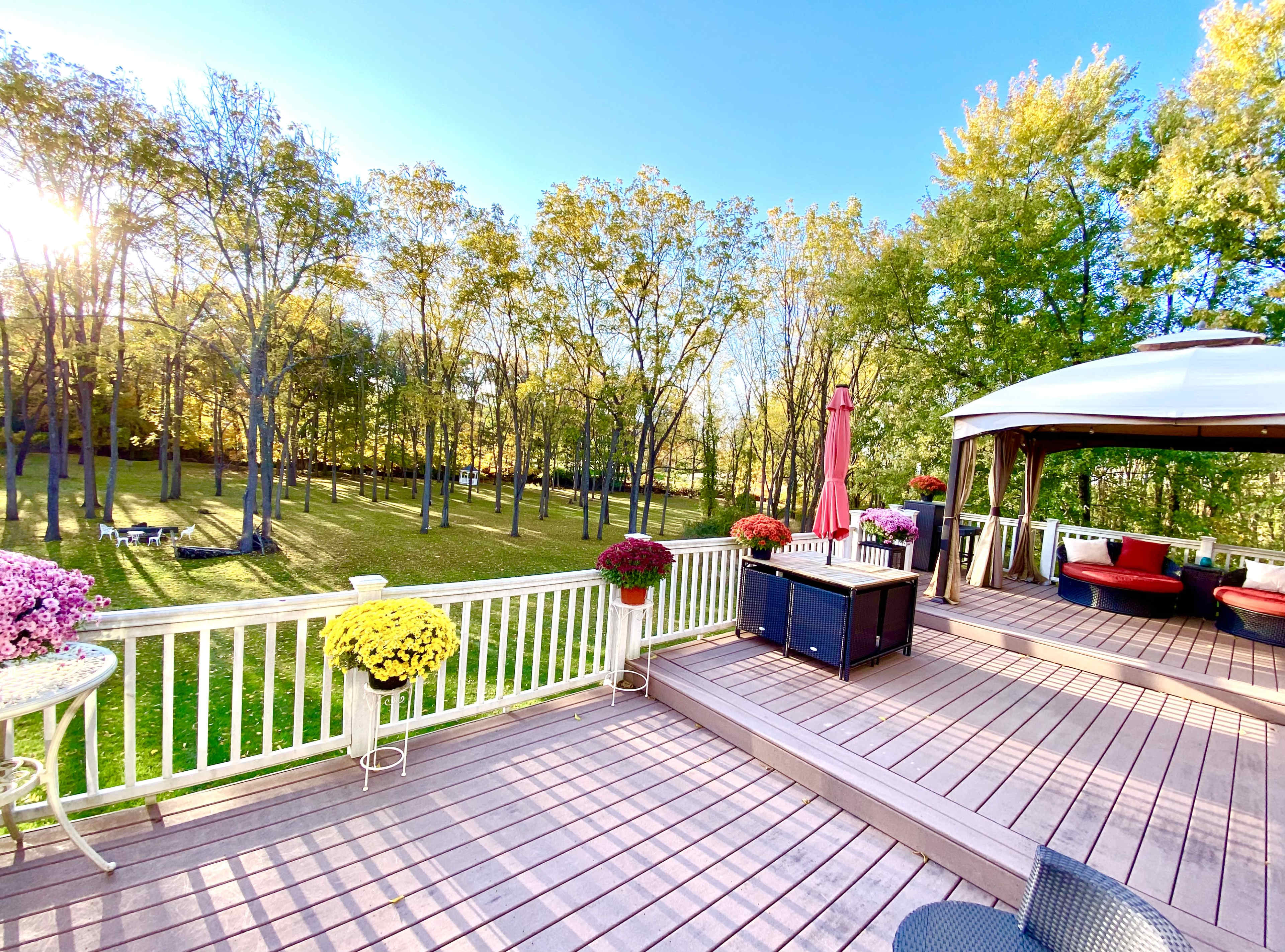 A well-furnished wooden deck is adorned with colorful potted flowers, overlooking a grassy area and a line of trees in the background under a clear blue sky.