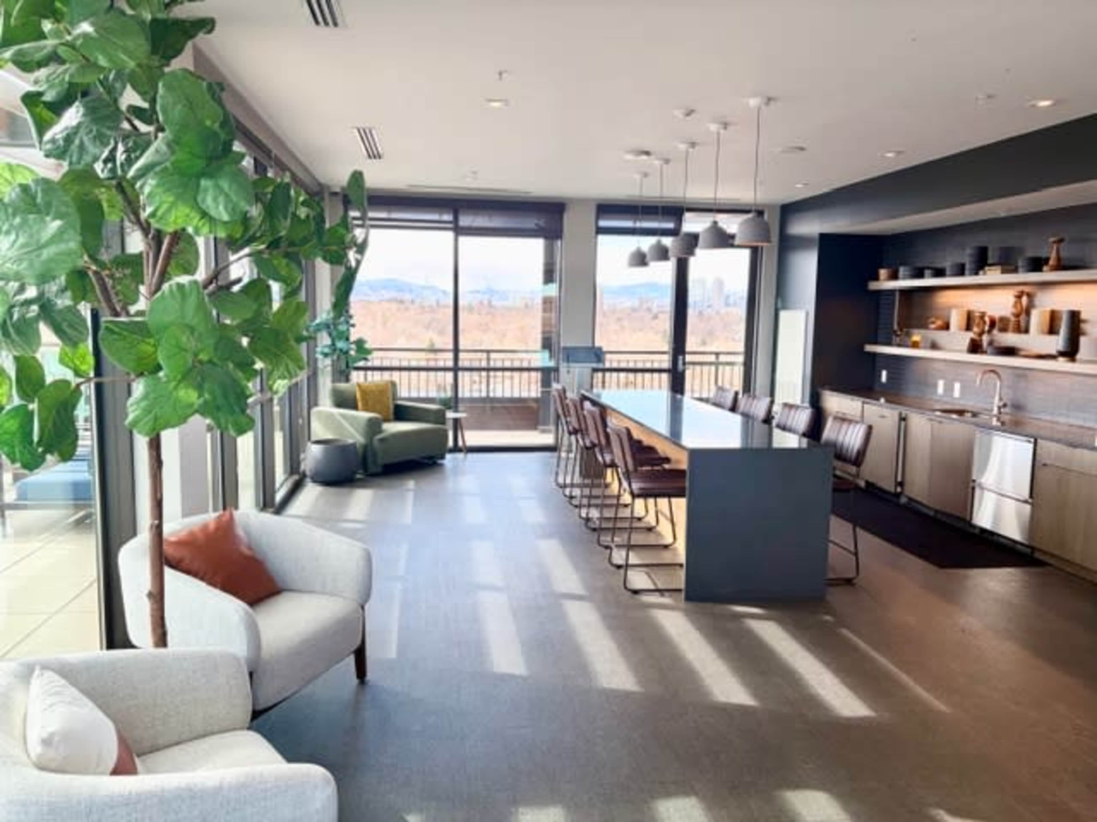 The image shows a modern, open-concept kitchen and dining area with large windows, featuring a long table, stylish chairs, and a greenery plant in the corner.