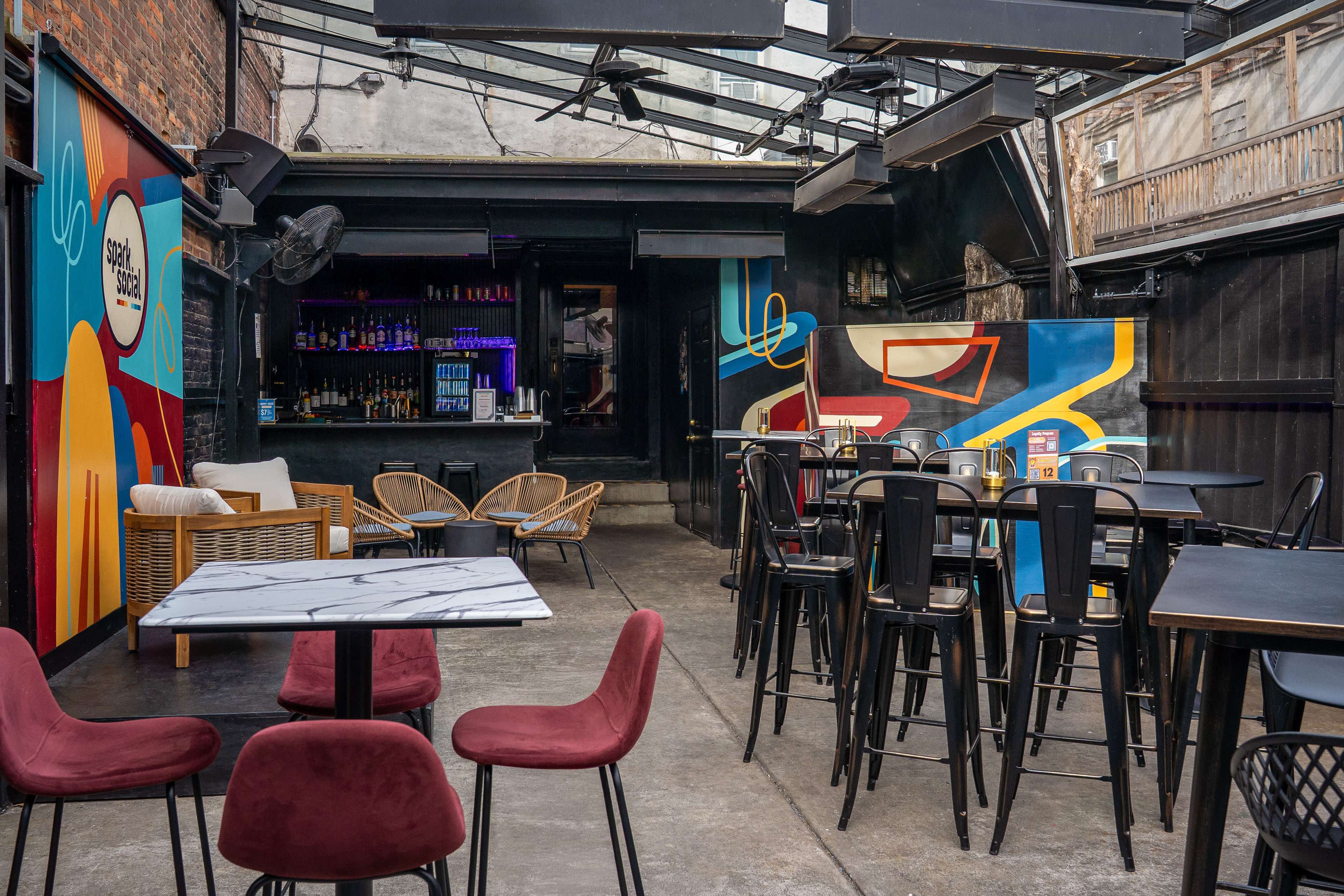The image shows an outdoor bar area with colorful murals, high tables, and chairs, and a bar counter in the background.