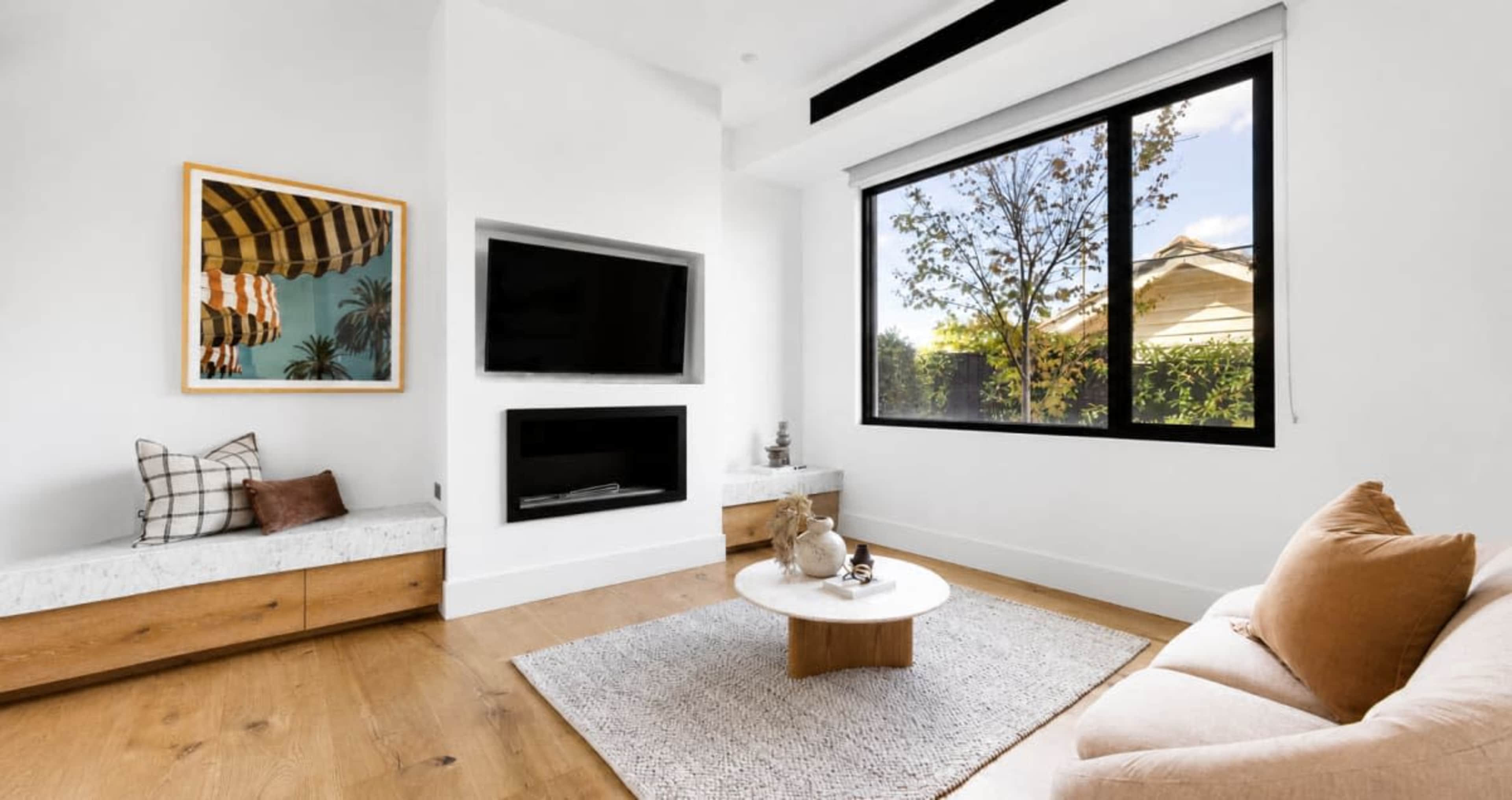 The image shows a modern living room with a large window, a mounted flat-screen TV, a light-colored sofa, and wooden furniture.