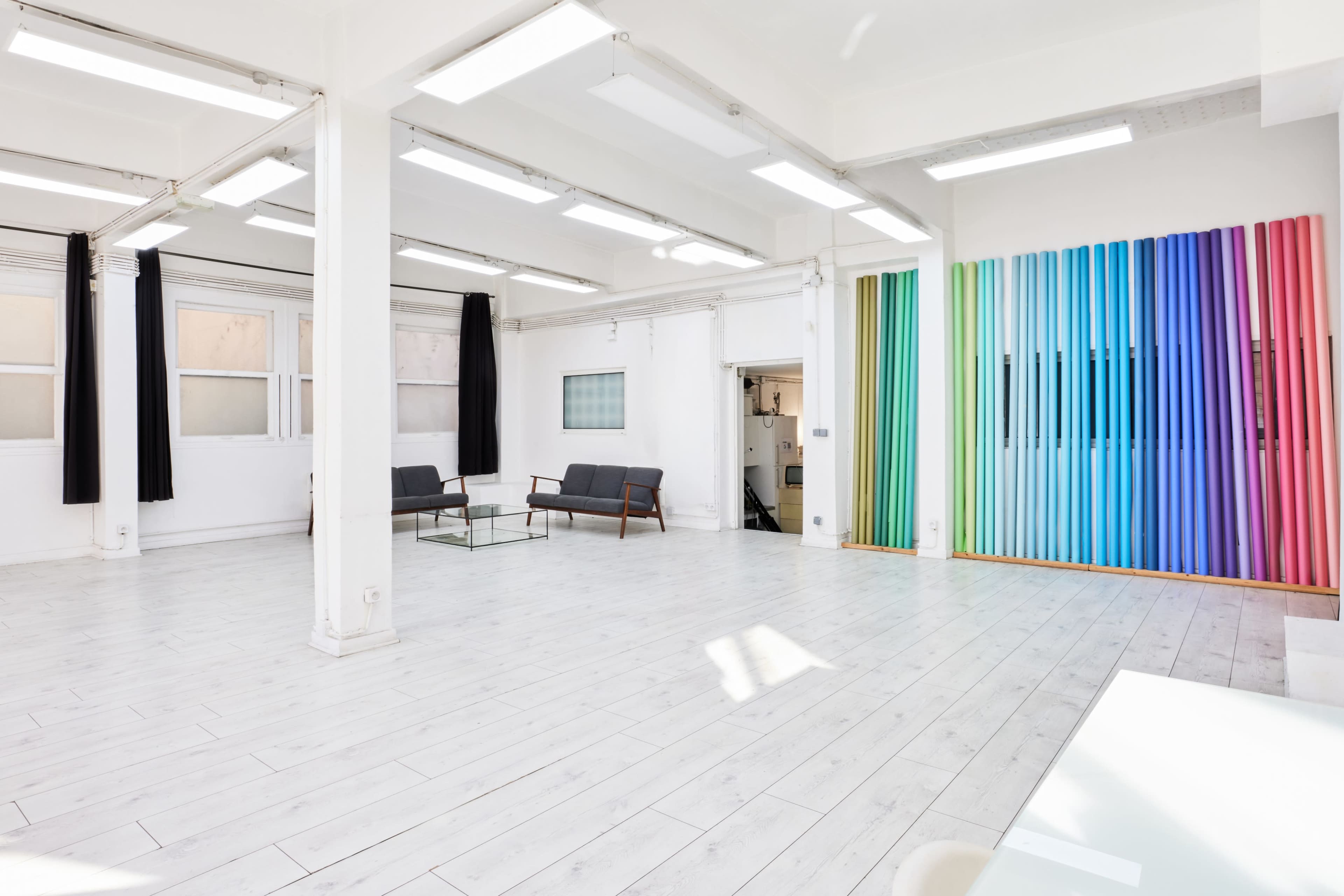 The image shows a spacious, well-lit studio with white walls, wooden flooring, and colorful vertical panels in one corner, along with a glass table and a gray sofa.