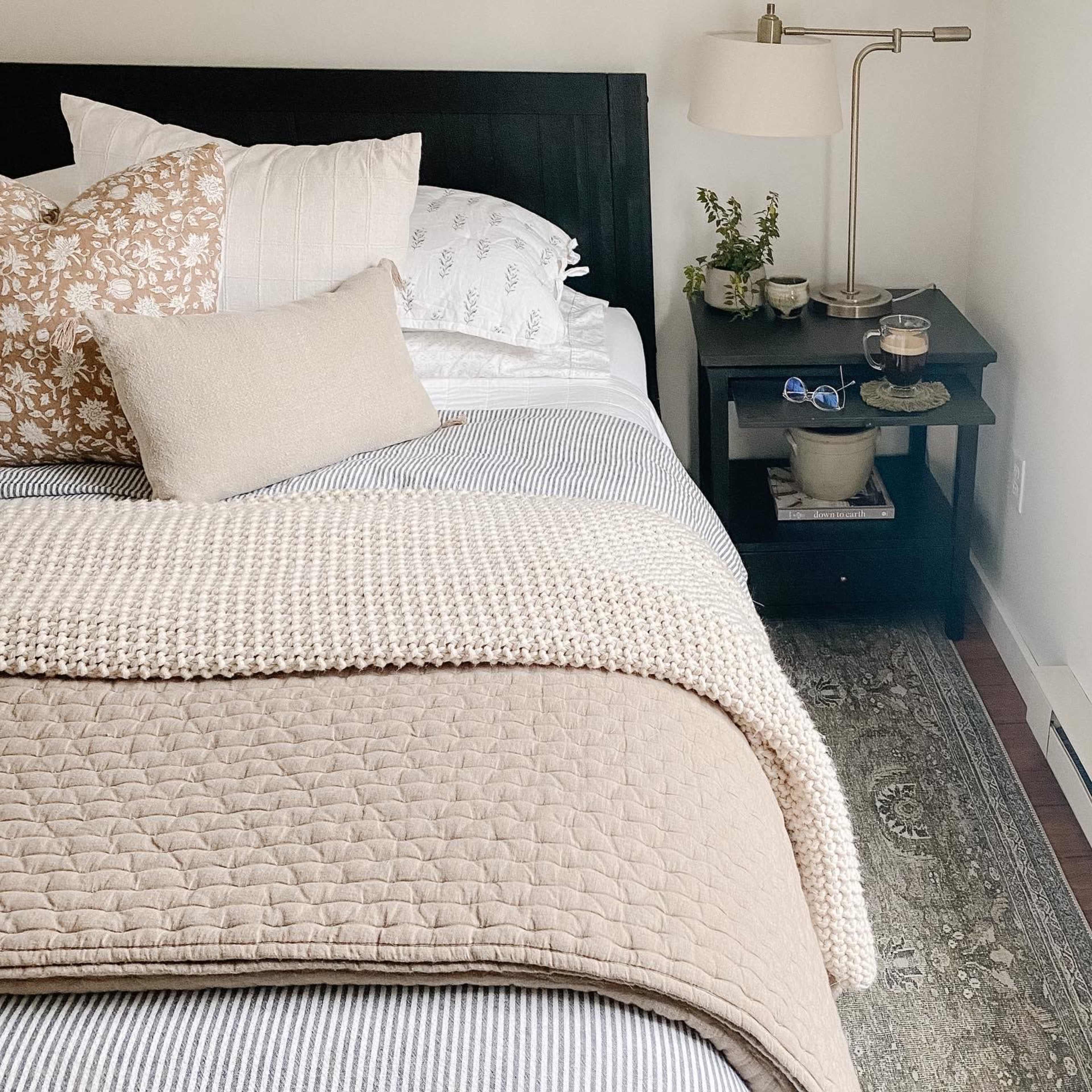 A neatly arranged bed features a striped blanket, decorative pillows, and is accompanied by a nightstand holding a plant and decorative items.