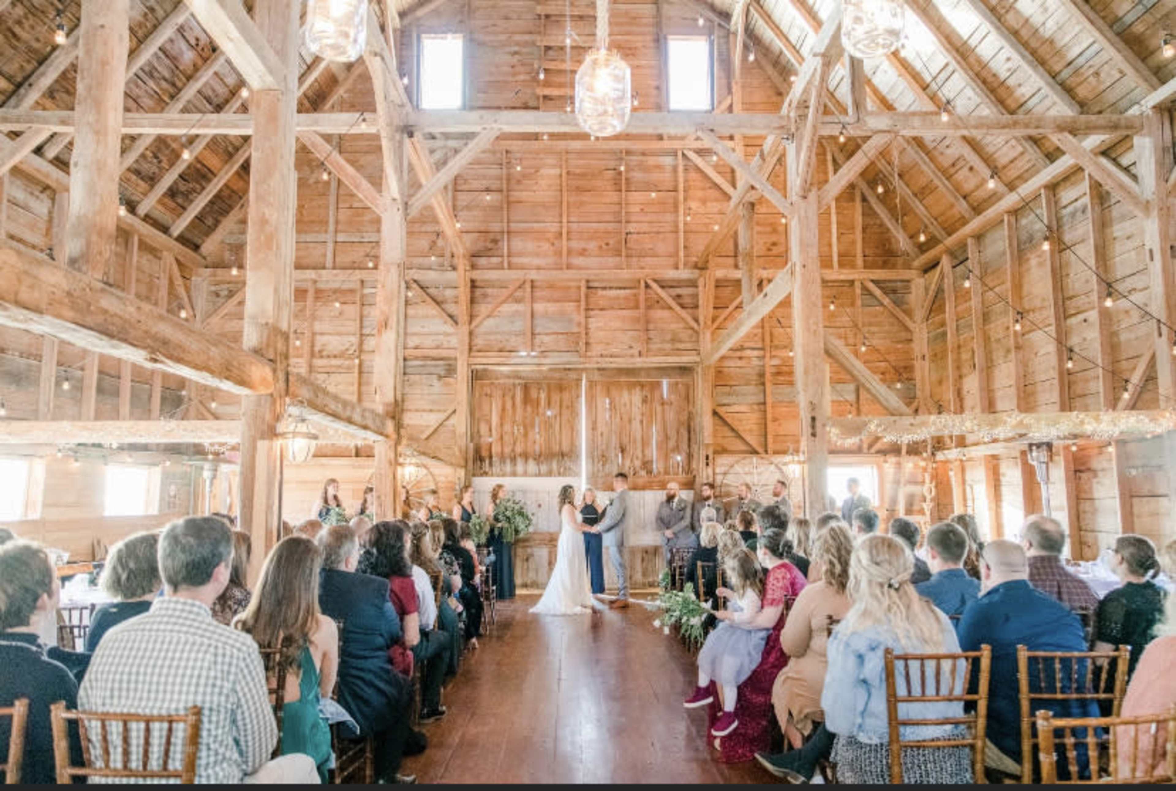 A couple stands at the front of a rustic barn, exchanging vows in front of a gathered audience of wedding guests.