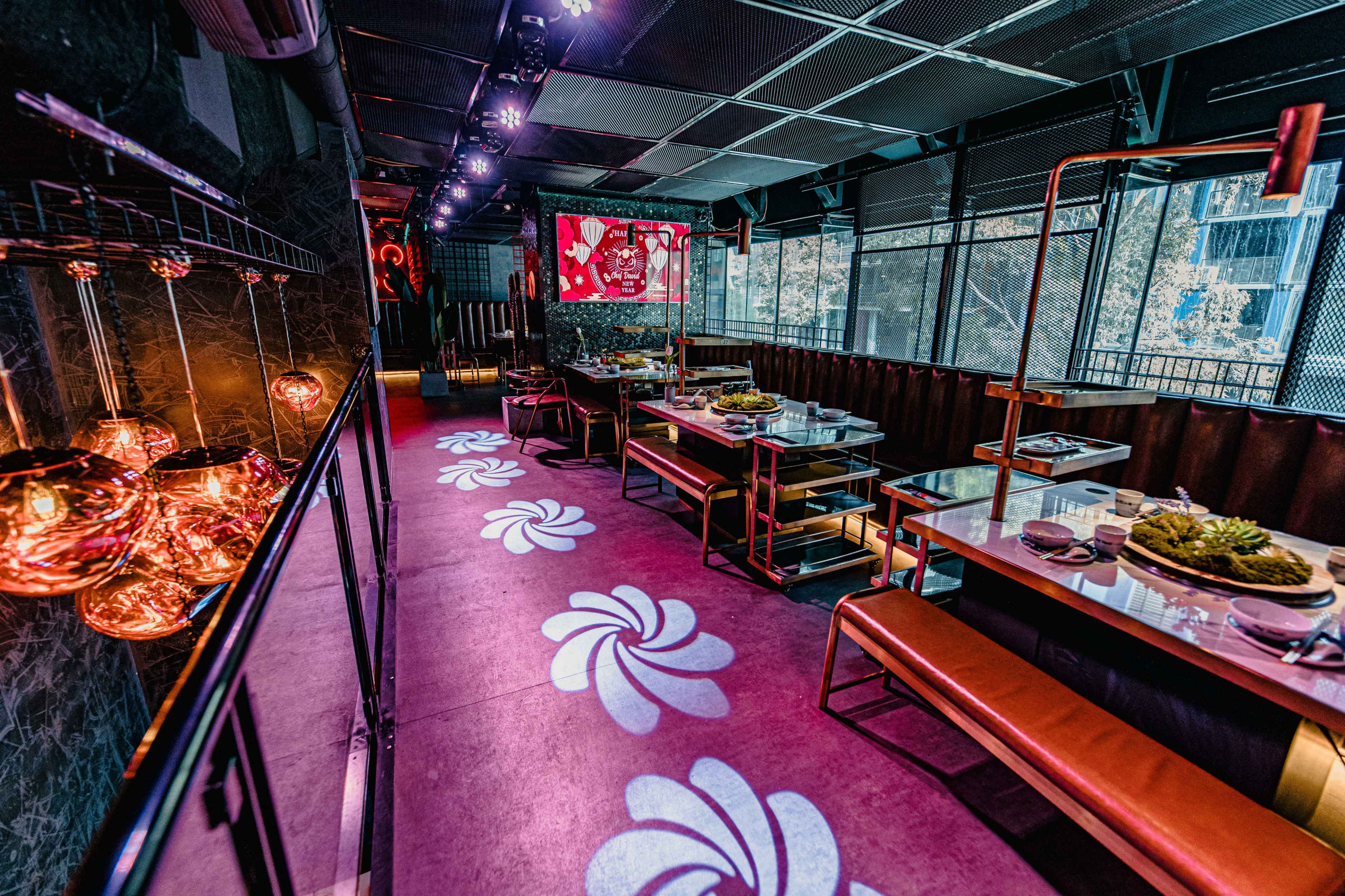 A dimly lit restaurant interior with modern furnishings, featuring tables along the sides and decorative lighting above, along with floral patterns projected on the floor.