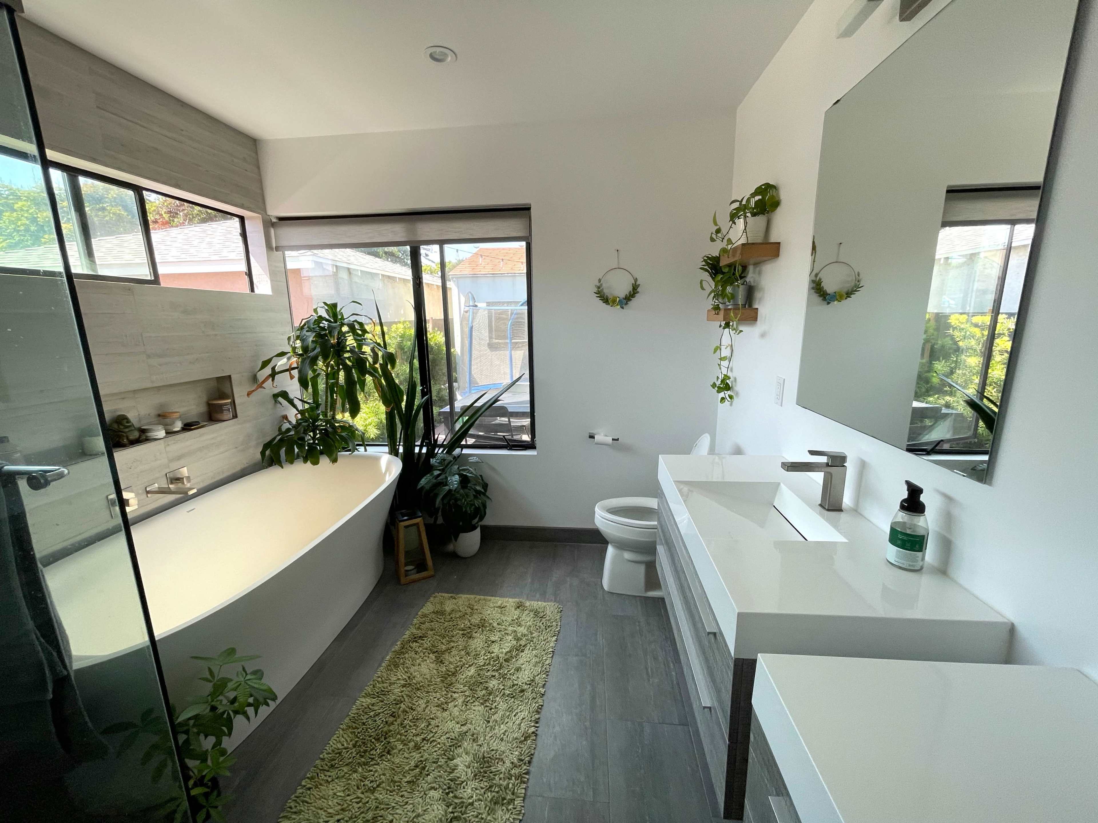 A modern bathroom featuring a freestanding bathtub, a large mirror above a vanity, and plants arranged around a window with natural light.