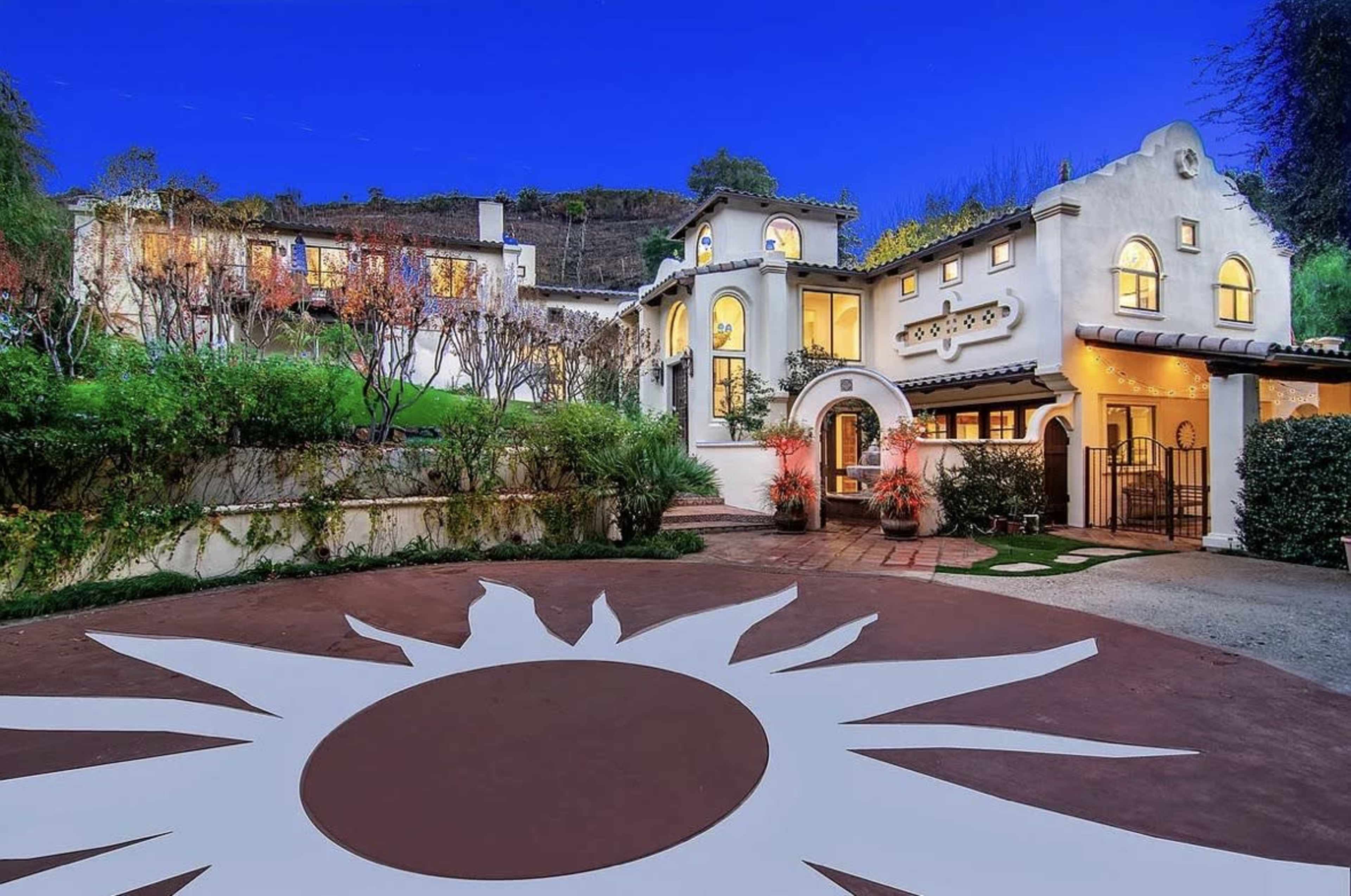 The image shows a spacious, two-story house with a decorative entrance and a circular sun pattern painted on the driveway, surrounded by landscaped gardens and trees under a twilight sky.