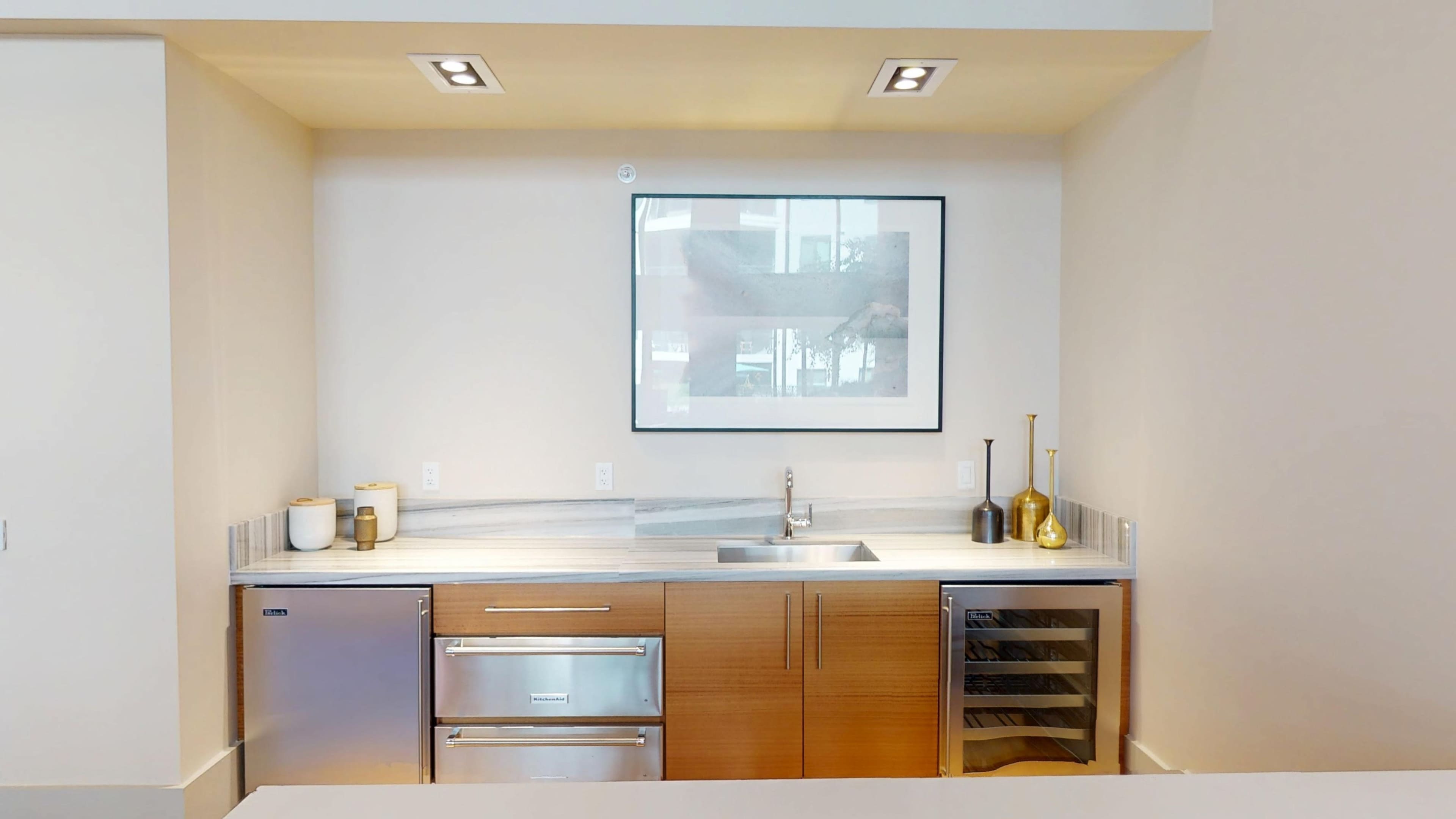 The image shows a modern kitchen featuring a stainless steel sink, wooden cabinetry, and a wine cooler alongside wall-mounted lighting and a framed artwork.