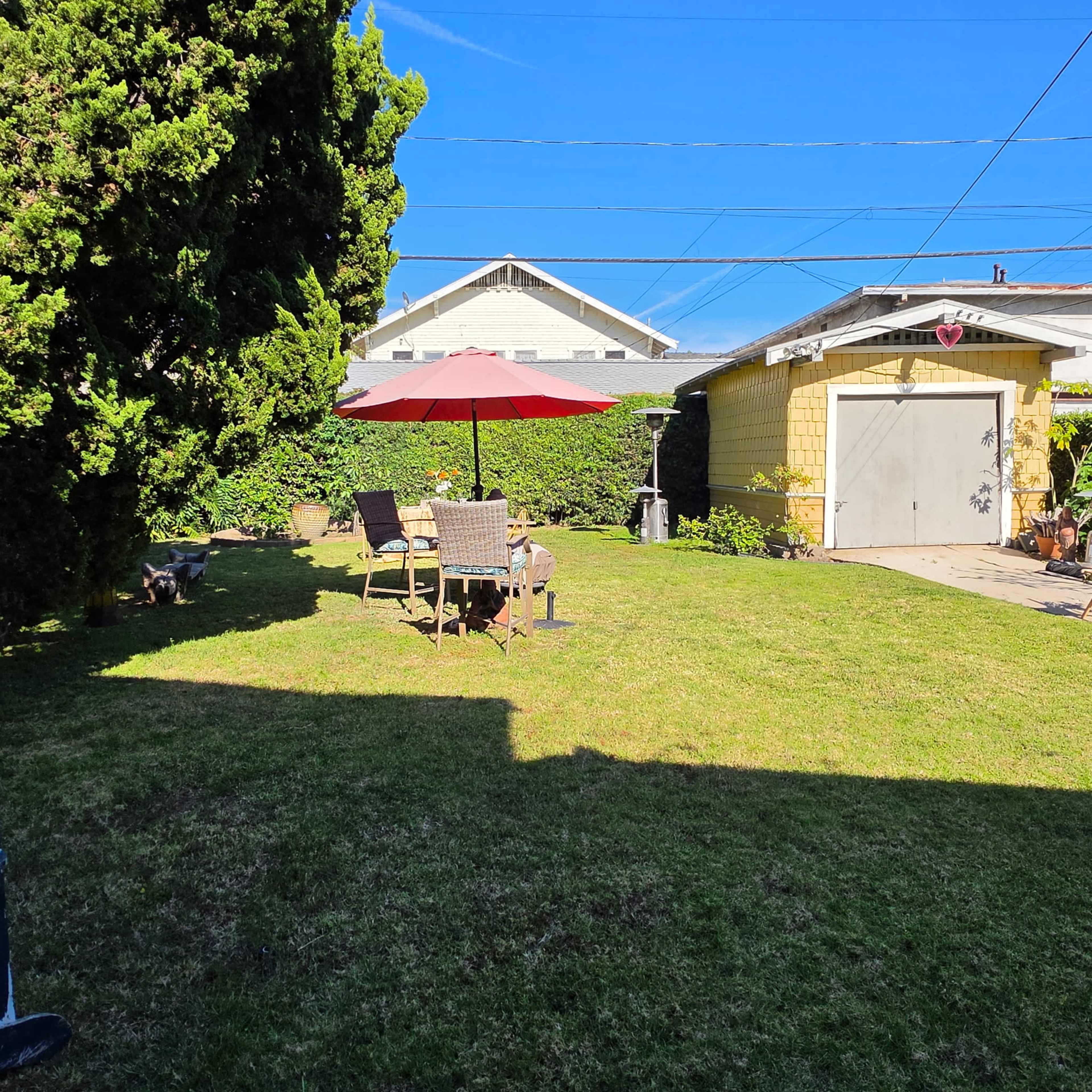 The image shows a grassy backyard with a patio set under a red umbrella, surrounded by shrubs and a garage in the background.