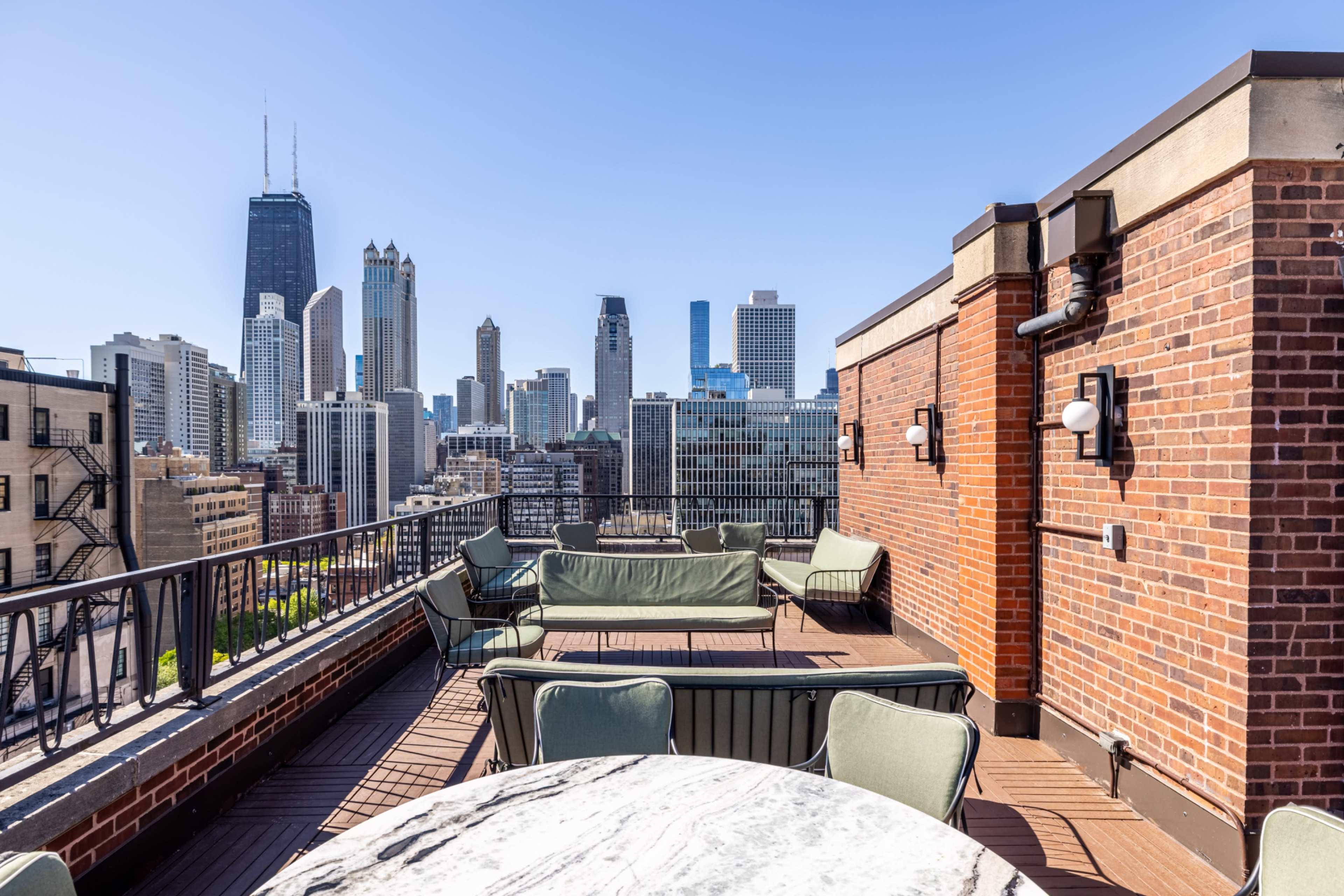 The image shows a rooftop terrace with seating and a view of skyscrapers in a city skyline under clear blue skies.