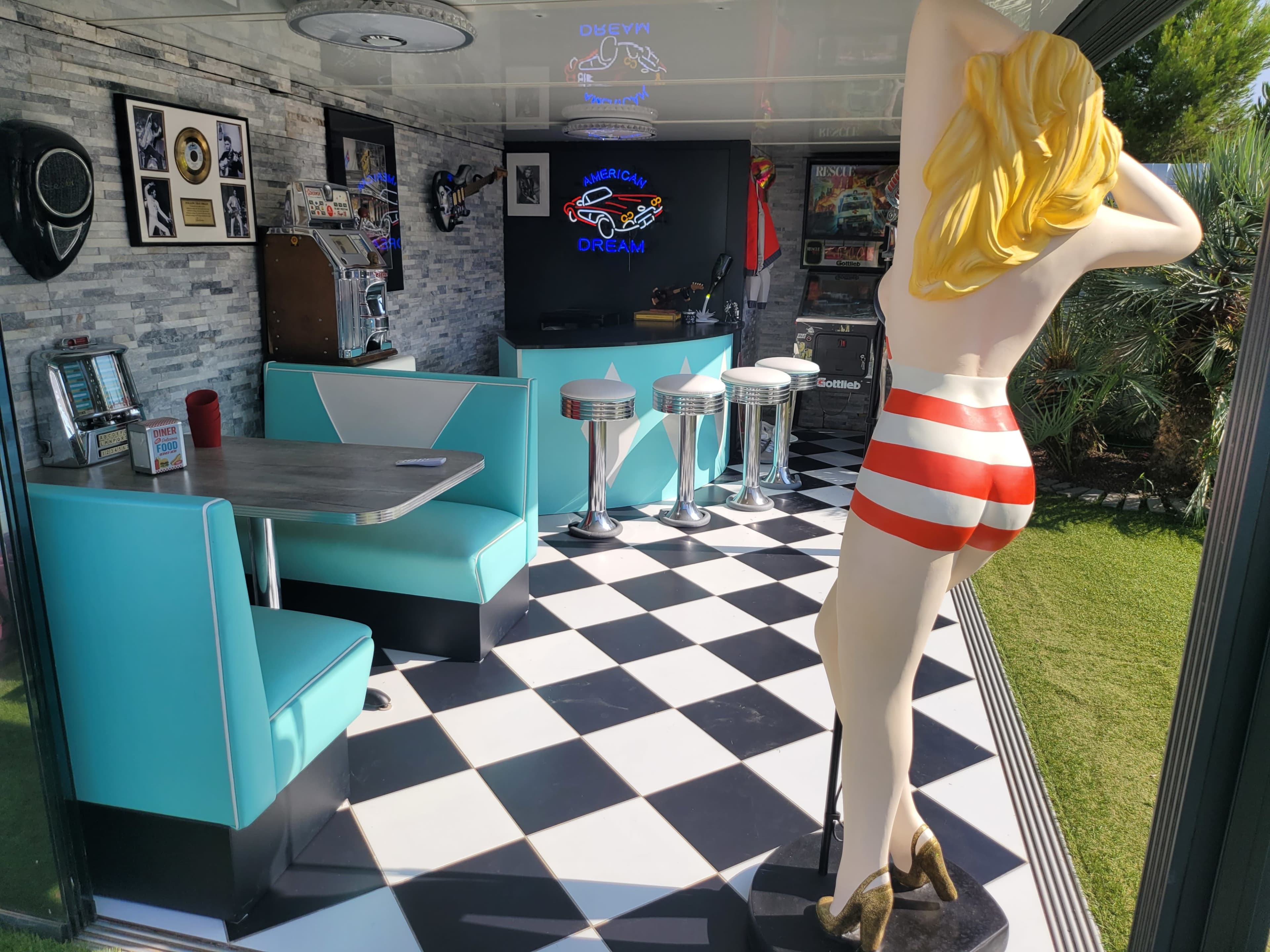 The image shows a retro-themed diner with aqua-colored seating, a black and white checkered floor, and a large statue of a woman in striped shorts standing by the entrance.