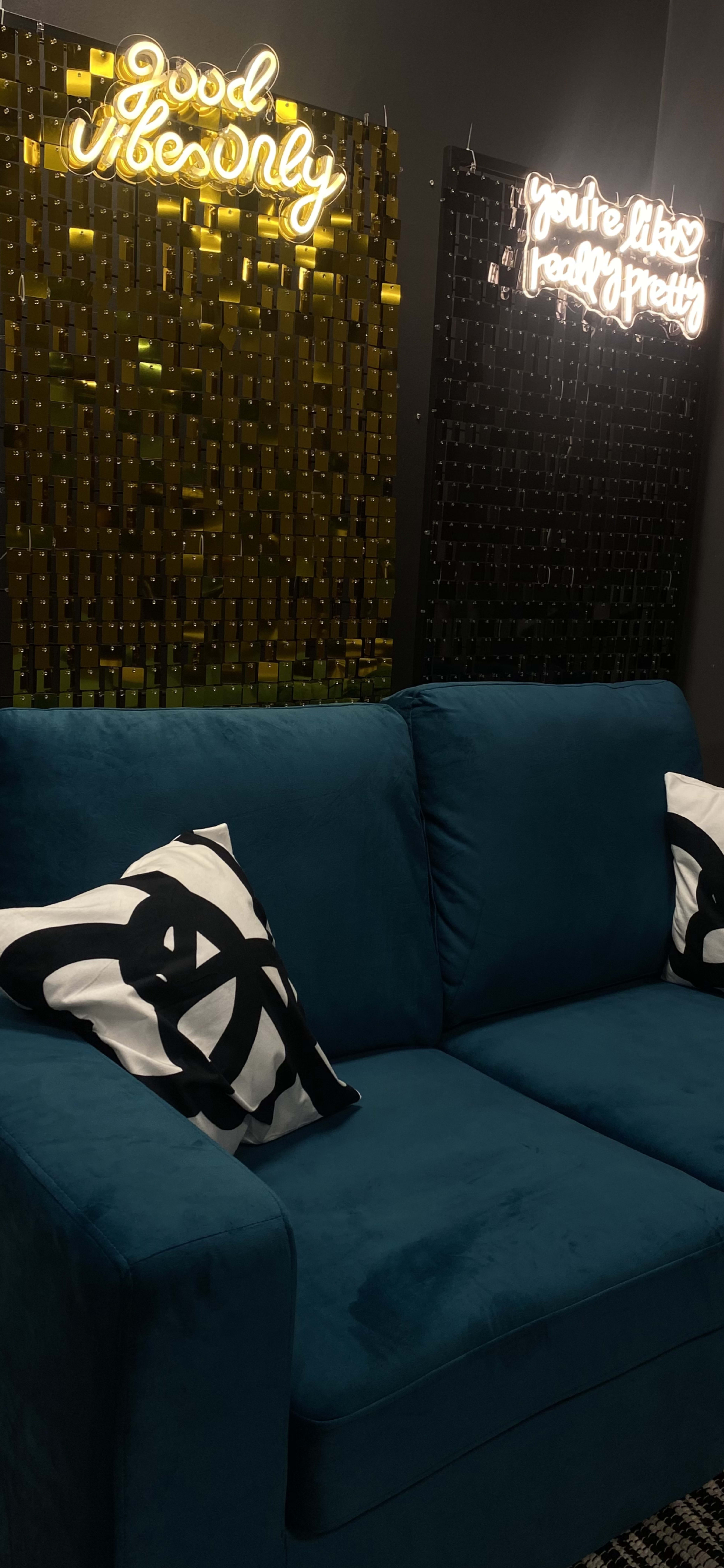A teal sofa with decorative pillows sits in front of two neon signs on a dark wall, one reading "Good Vibes Only" and the other "You're like really pretty."