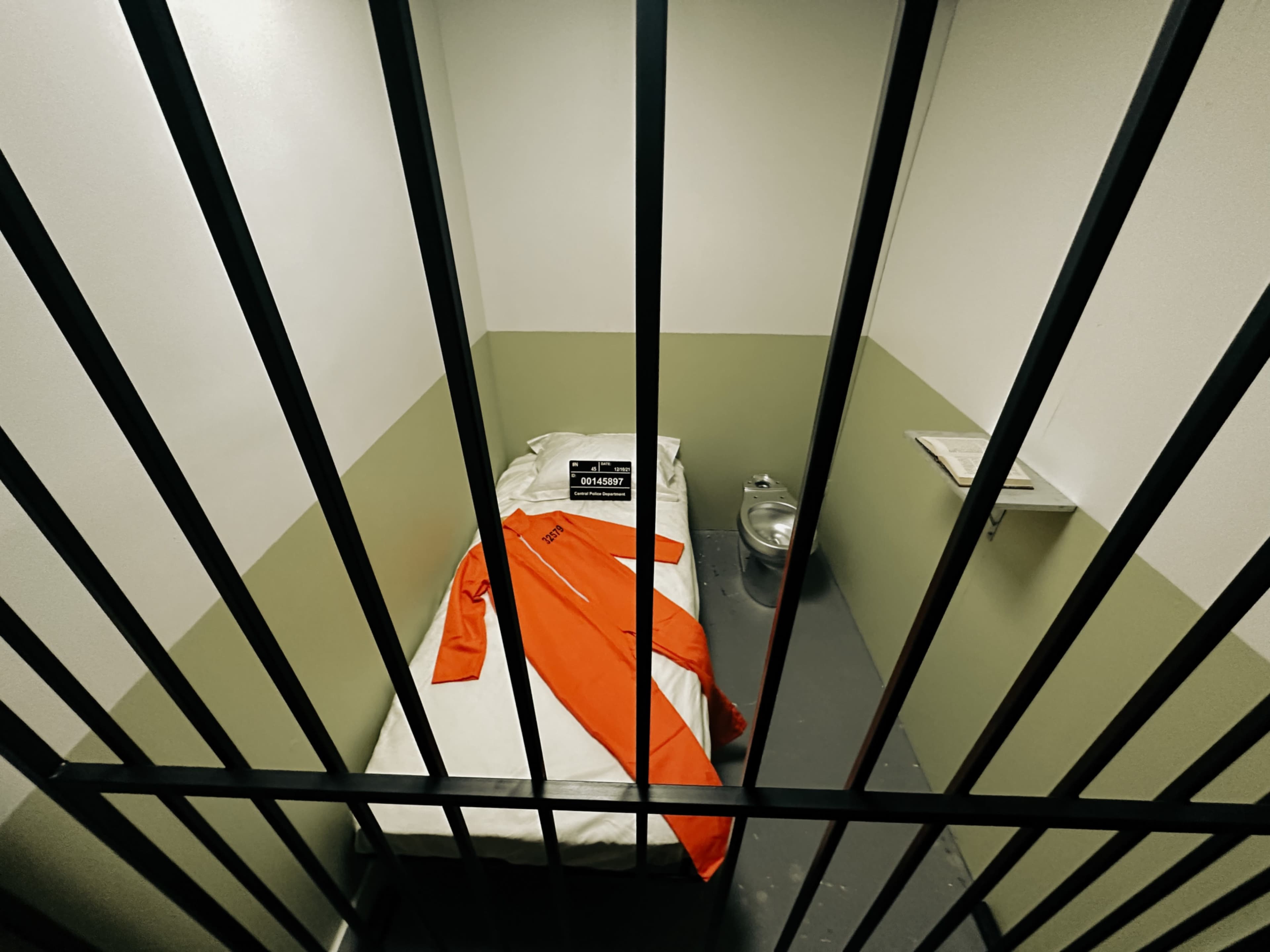 A small jail cell features a bed with white linens, an orange jumpsuit laid across it, a metal toilet, and a window with bars.