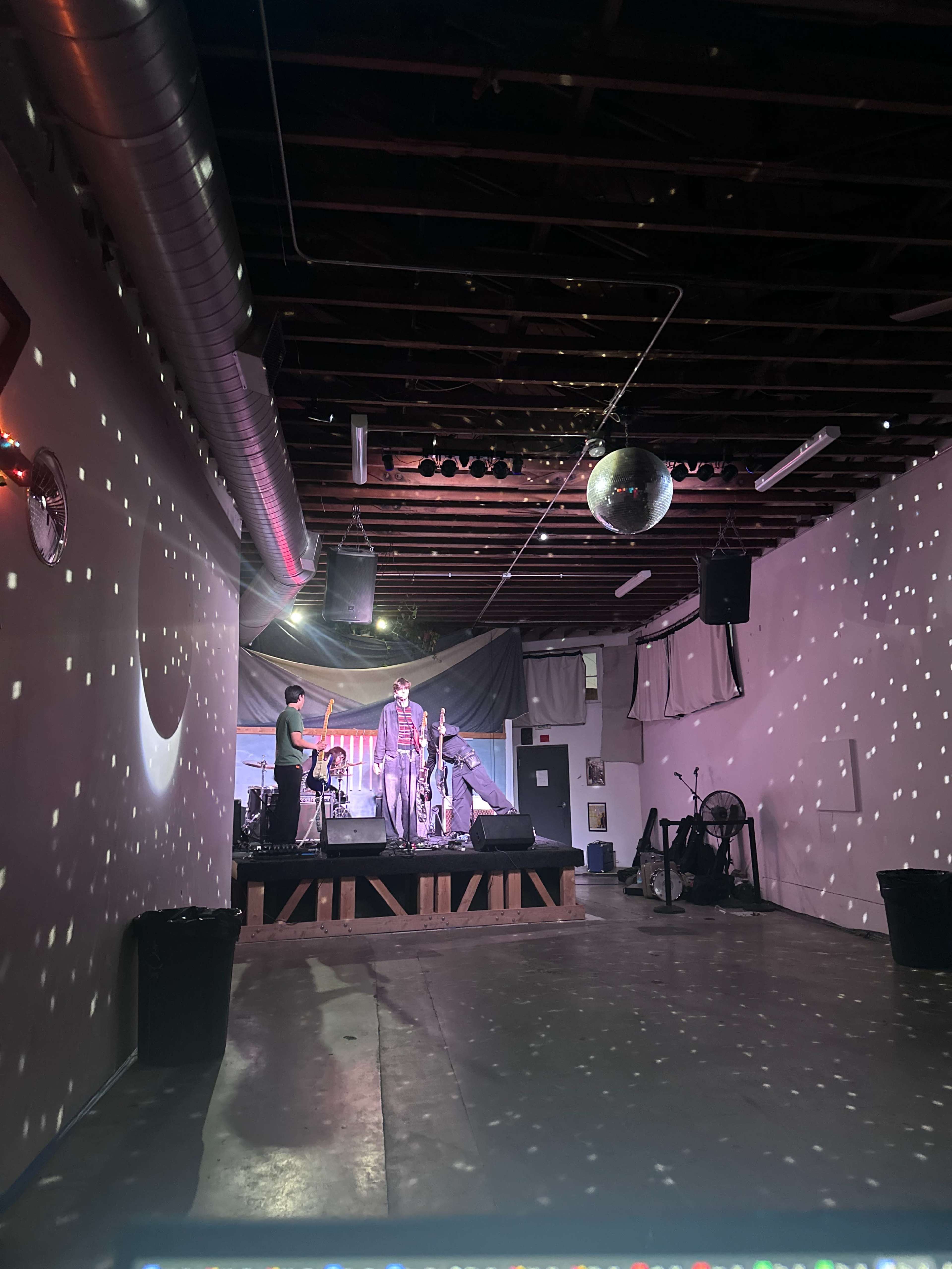 The image shows a performance space with a stage, drum set, and a person standing at the front, illuminated by colorful lights and a large disco ball reflecting patterns on the walls.