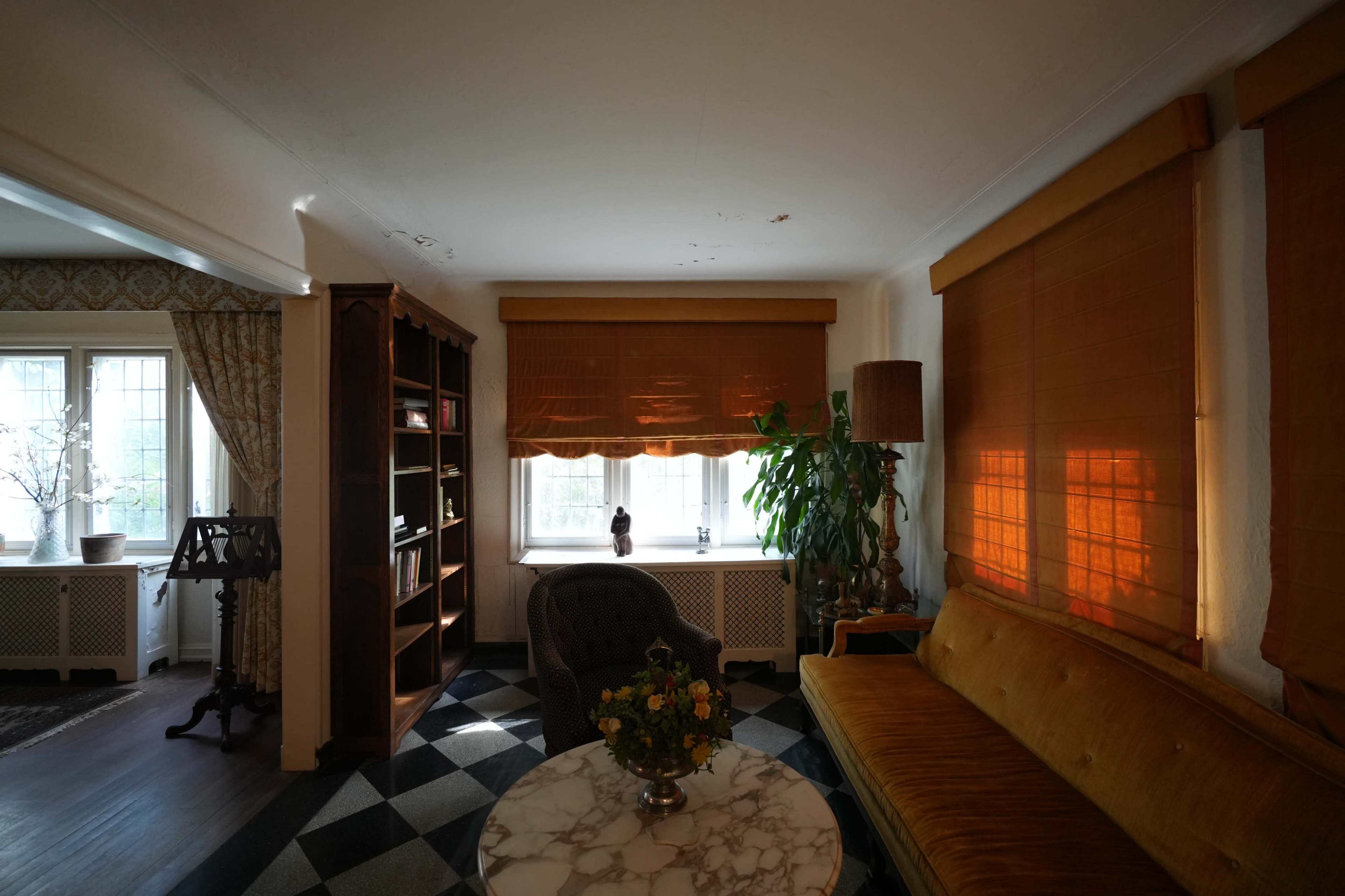 The room features a cozy seating area with a velvet sofa, a marble-topped table, and large windows dressed with wooden shades, illuminated by soft afternoon light.