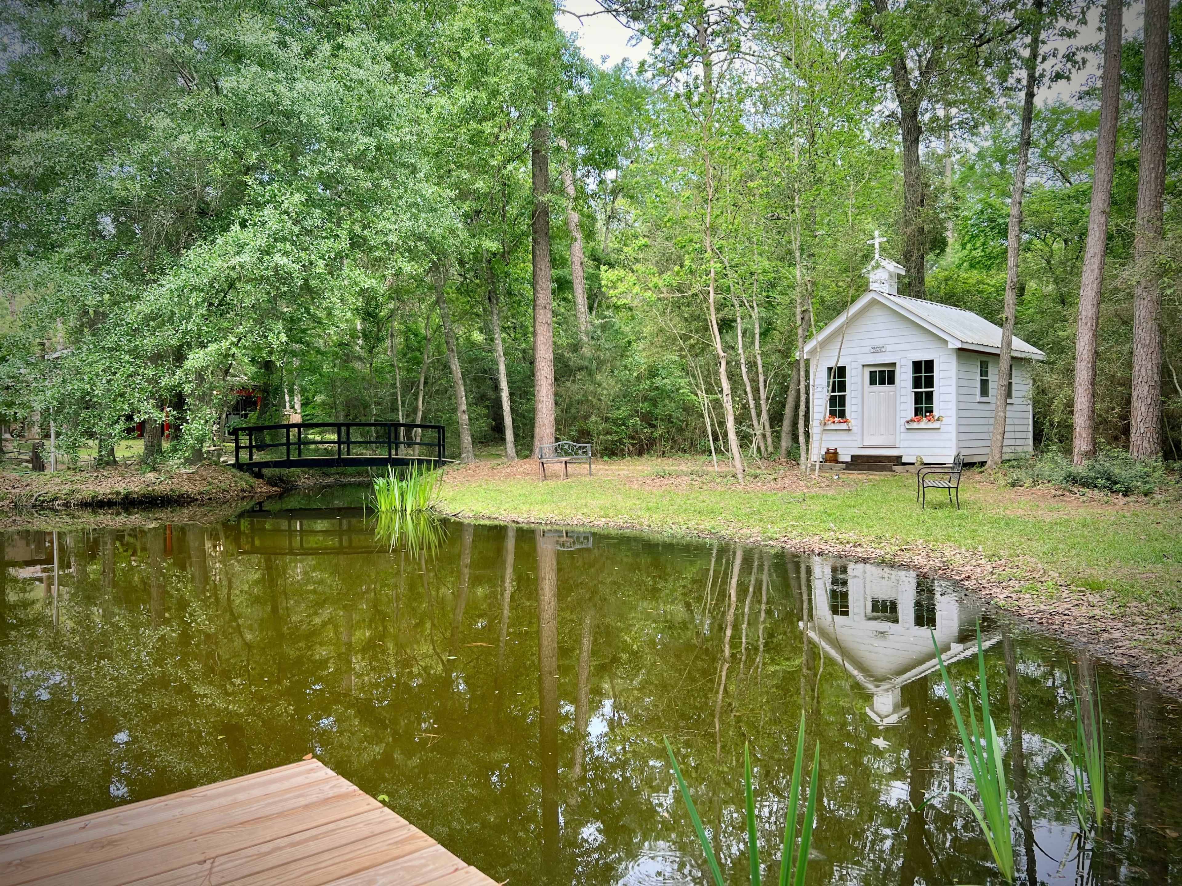 A small, white cabin with flower boxes stands by a pond surrounded by trees, with a wooden bridge visible in the background.