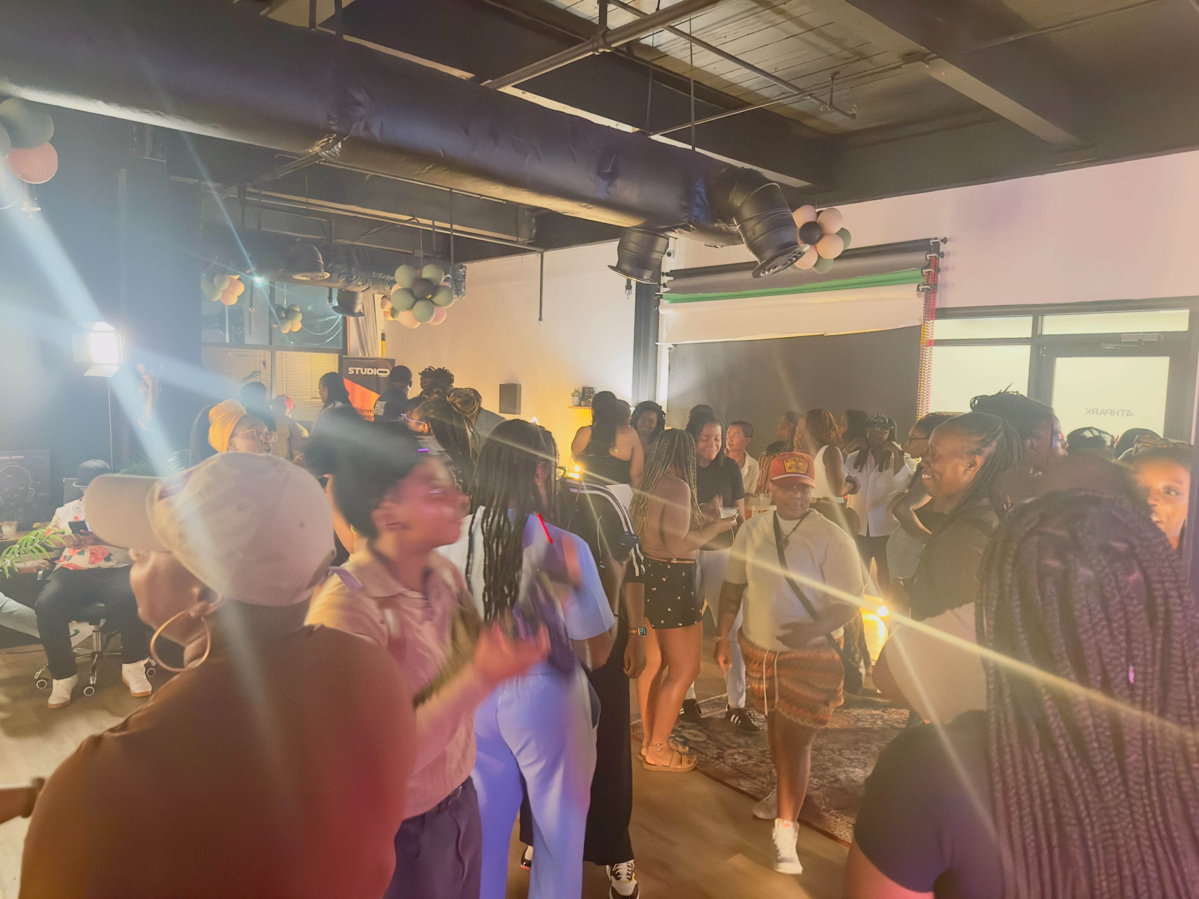 A lively indoor gathering features a diverse group of people socializing and dancing amidst colorful decorations.