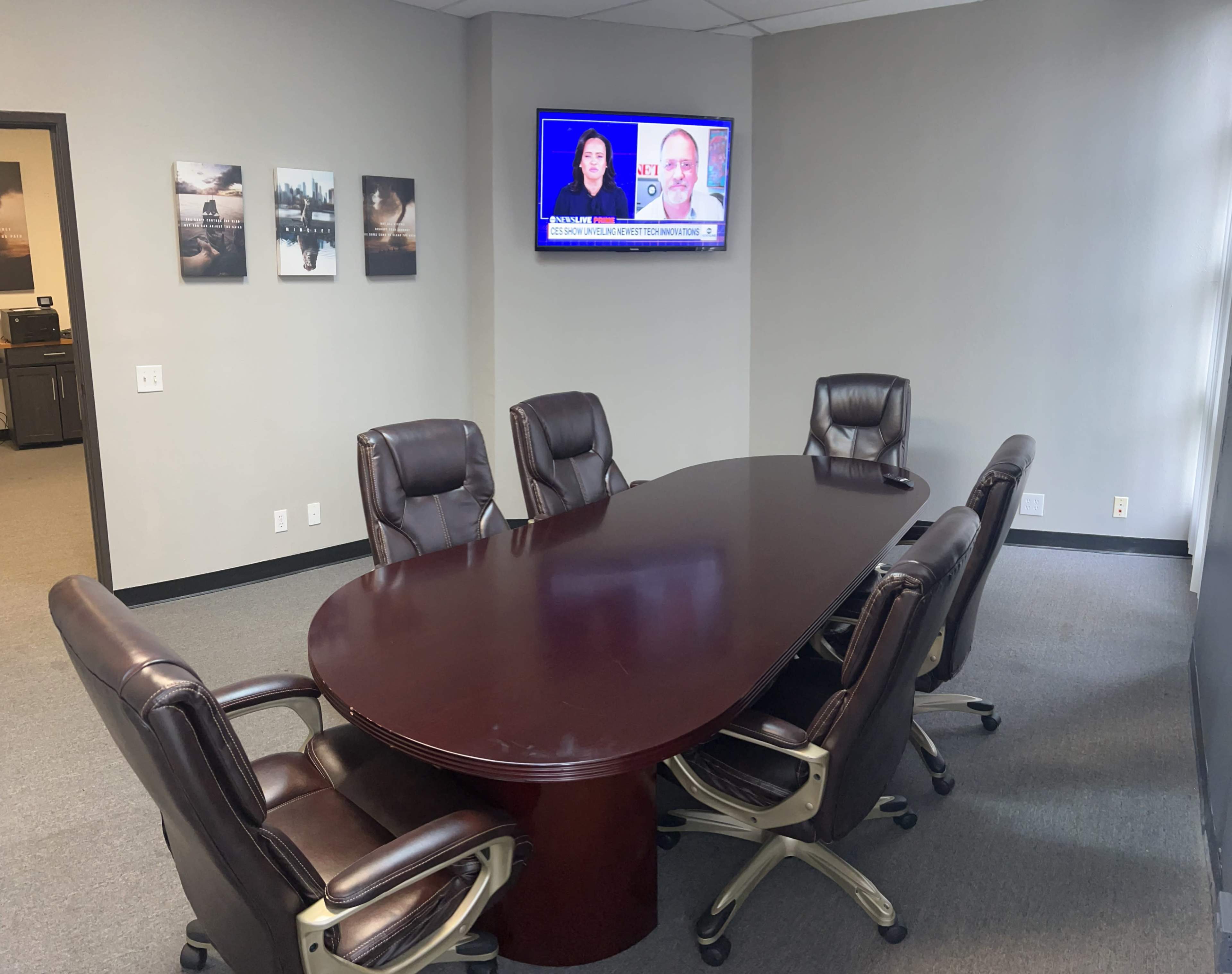 A conference room featuring a large oval table surrounded by six leather chairs, with a television displaying a news broadcast on the wall.
