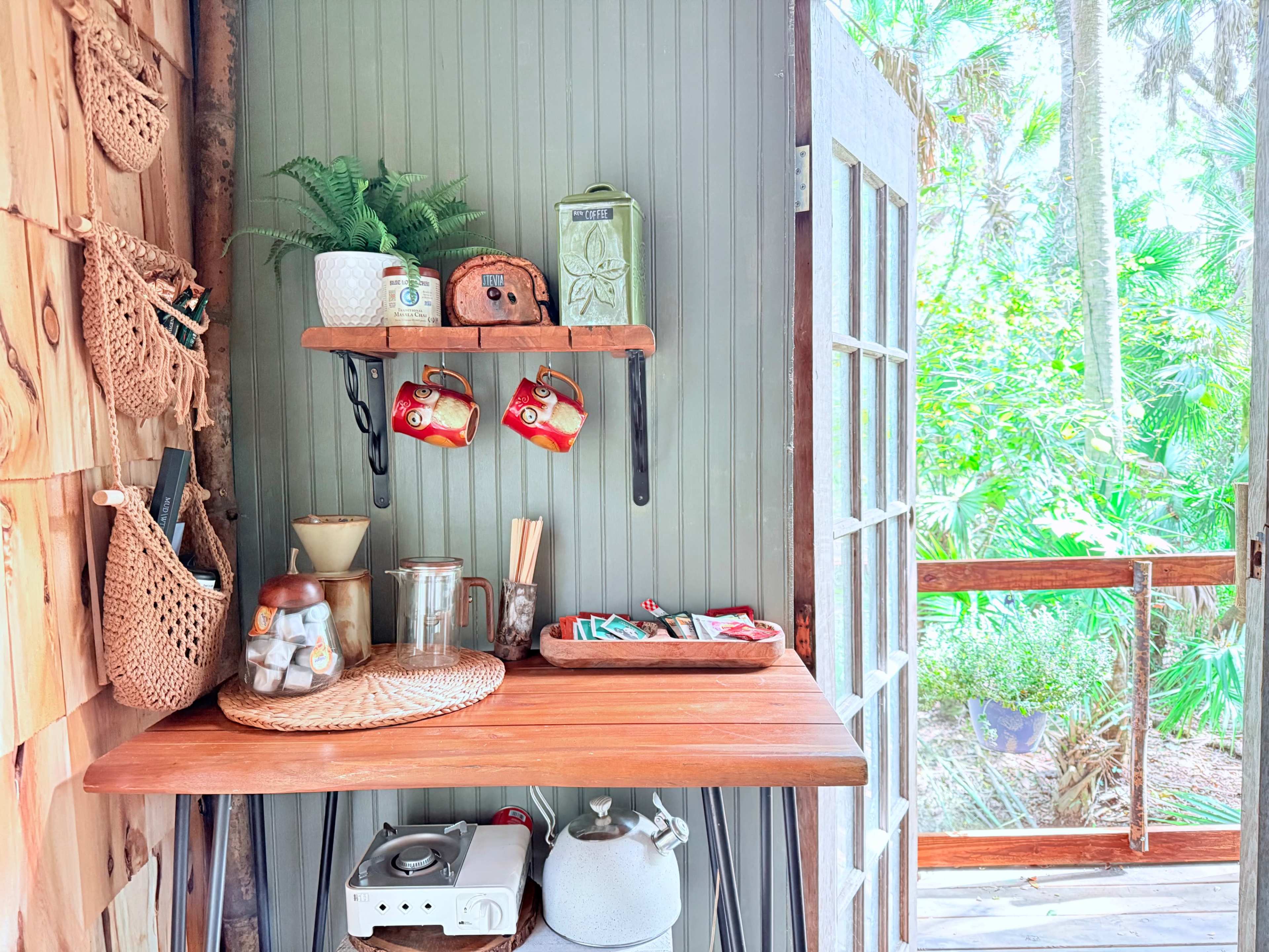 The scene shows a cozy outdoor kitchen area with wooden shelves displaying dishes and utensils, next to a door leading to a lush garden.